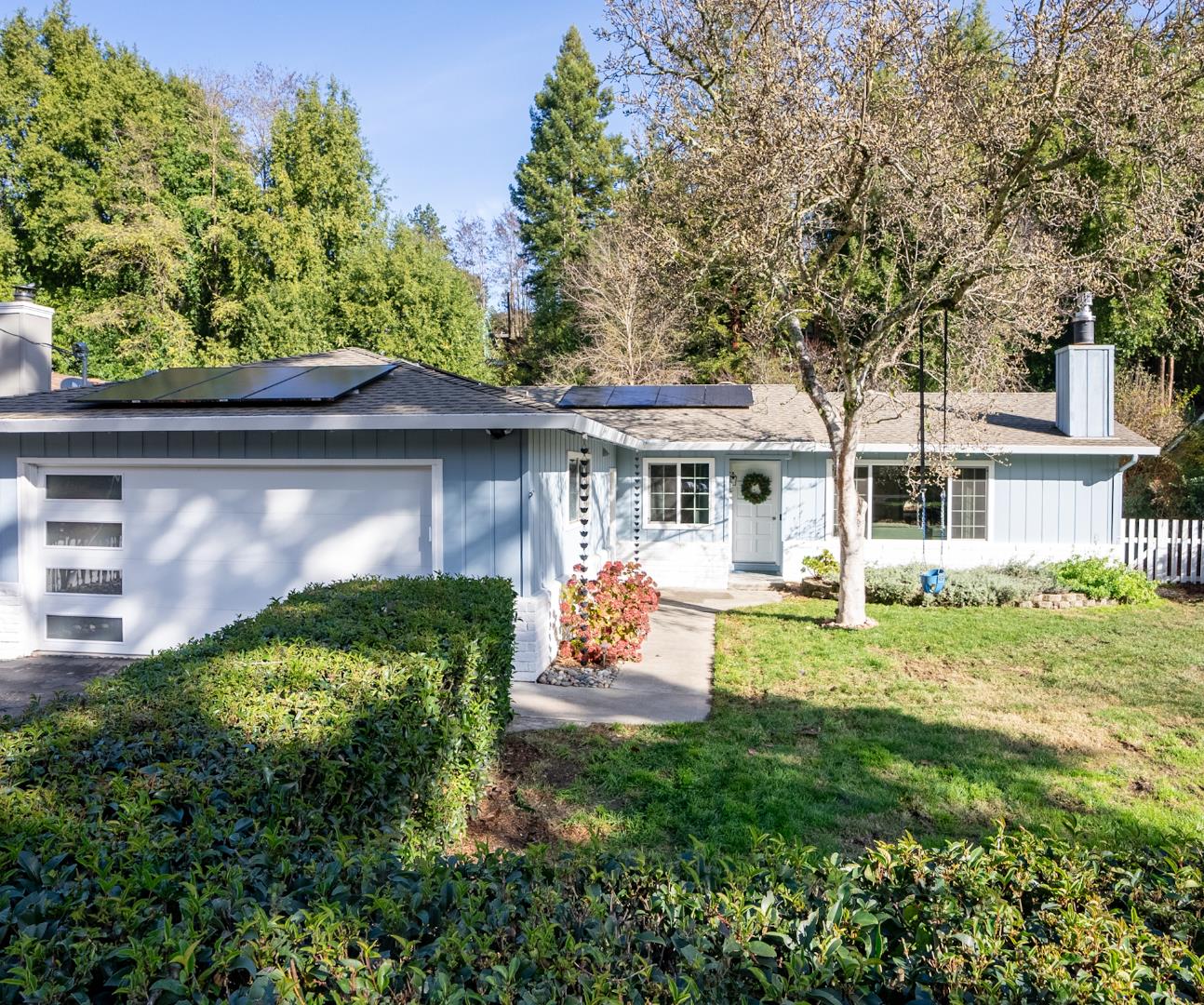 Single-story blue home with rooftop solar panels, white garage door, and landscaped front yard.