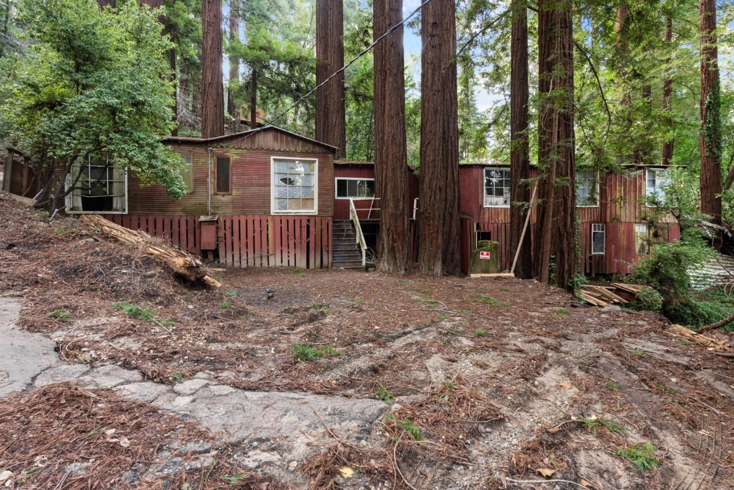 Weathered red house among towering redwood trees, with a rough clearing in front.