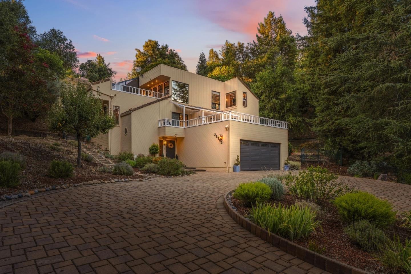 Multi-level beige home with balconies, a dark garage, and a curved paver driveway at dusk.