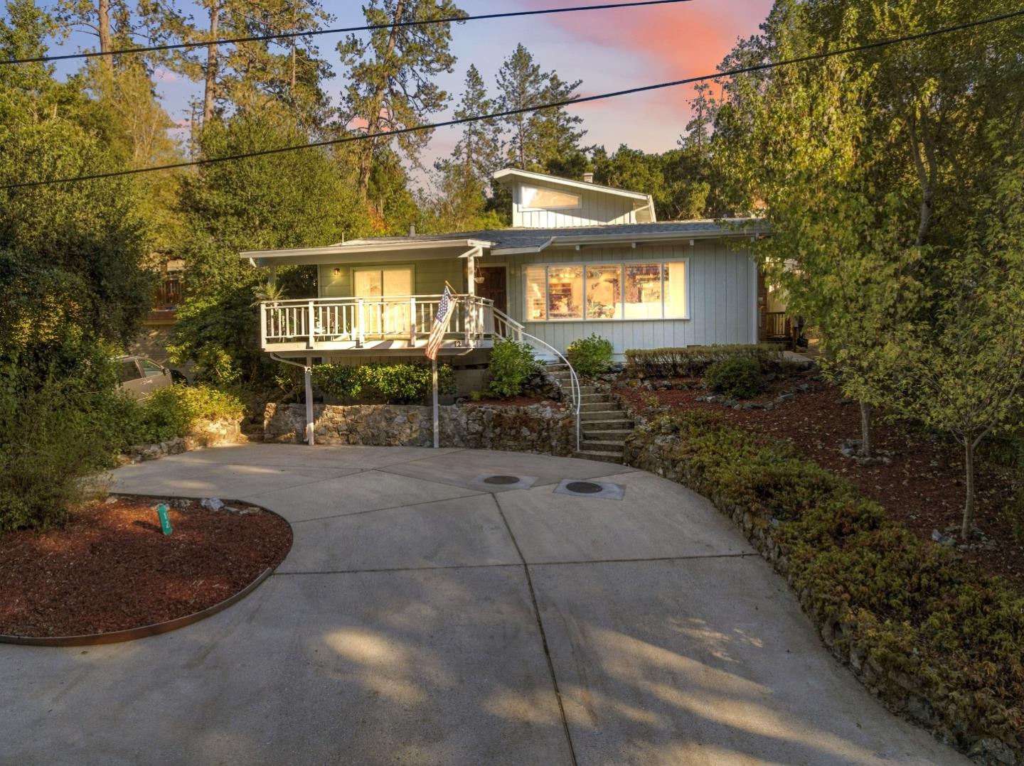 Gray home with a raised front deck and American flag, set among mature trees beside a curved driveway.