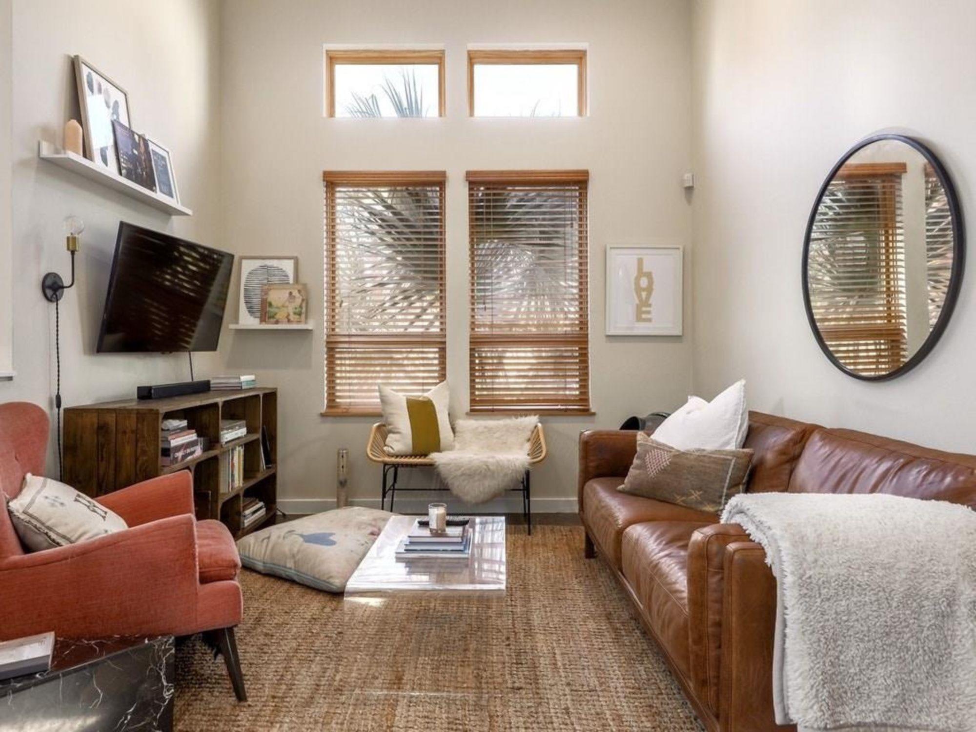Furnished living room with a brown leather sofa, coral armchair, and tall windows with wooden blinds.