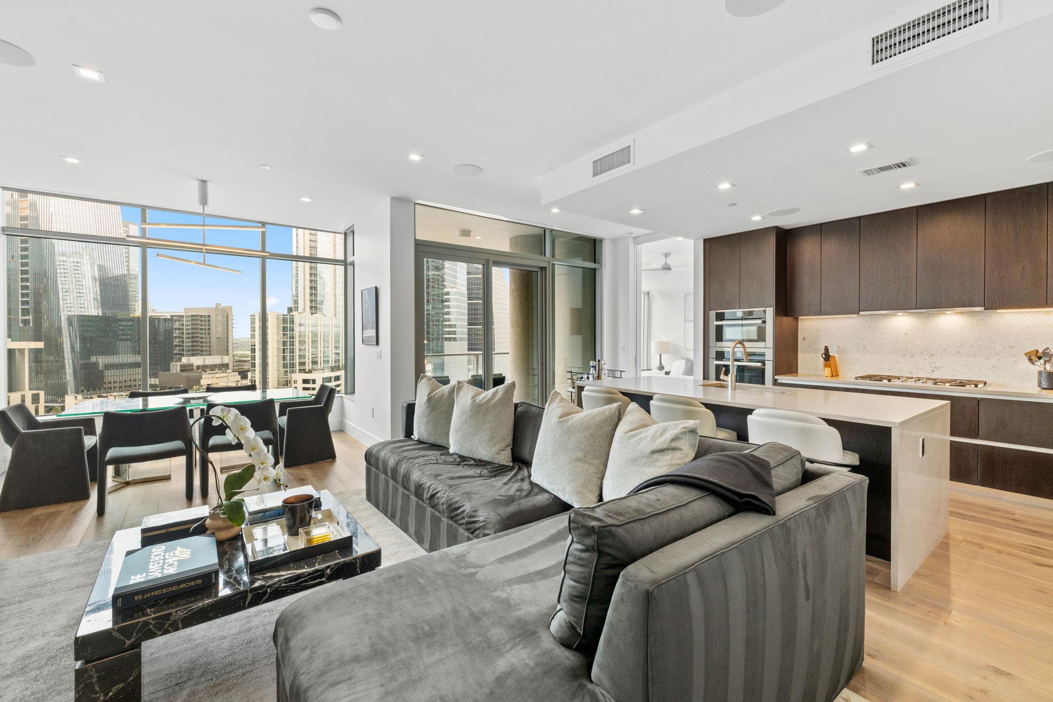 Open-plan condo living room with a gray sectional, modern kitchen island, and city views through floor-to-ceiling windows.