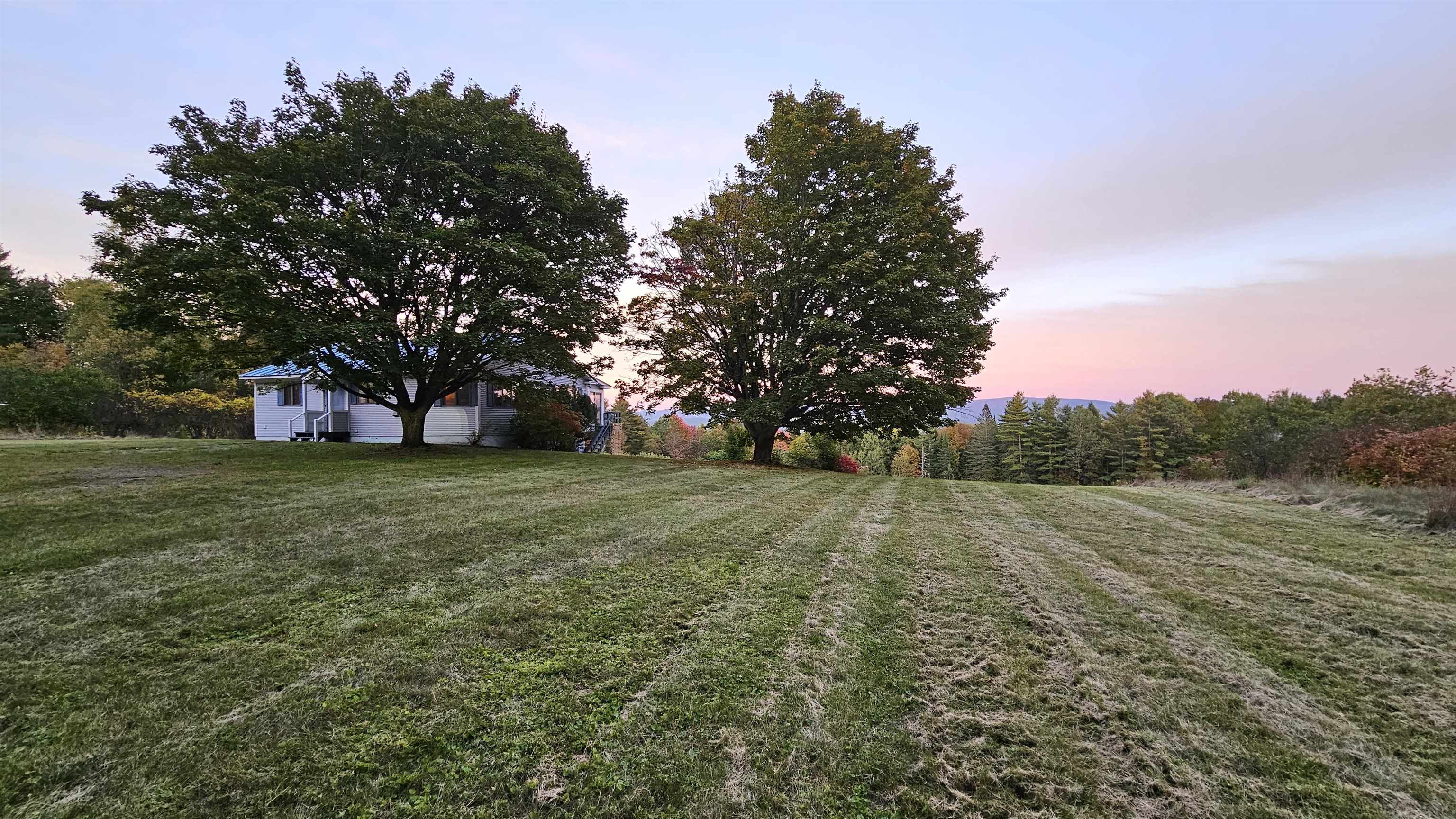 White house partly hidden by mature trees above a broad lawn, with a wooded ridge under a pink dusk sky.