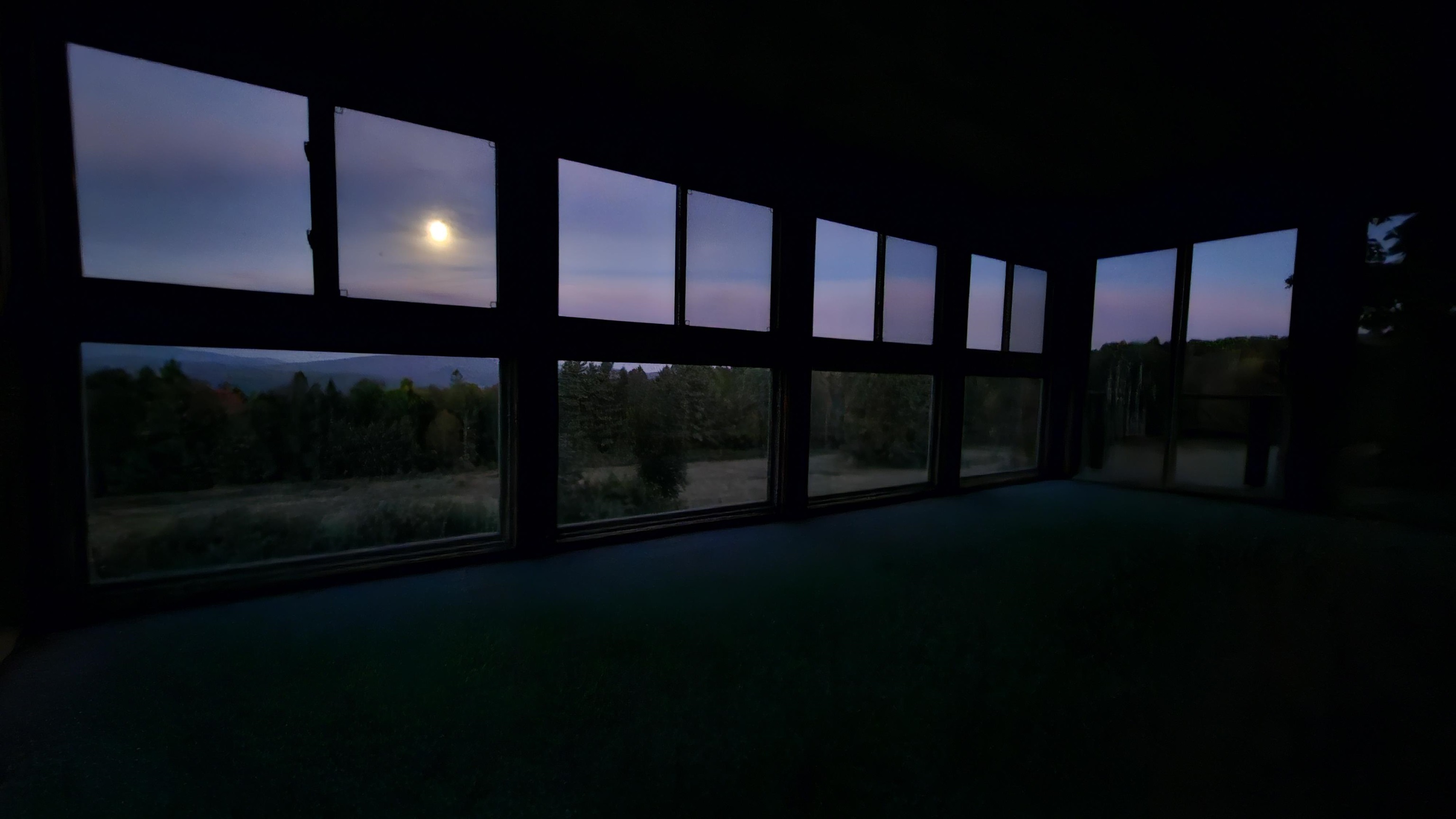 Vacant room with expansive windows framing distant hills and a bright moon at twilight.