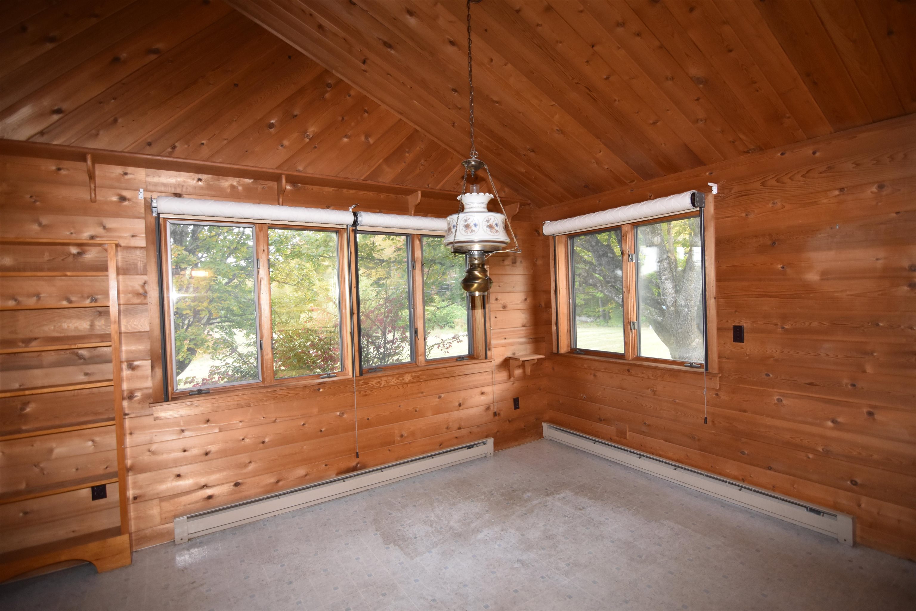 Empty pine-paneled room with vaulted ceiling, built-in shelves, and broad windows overlooking trees.