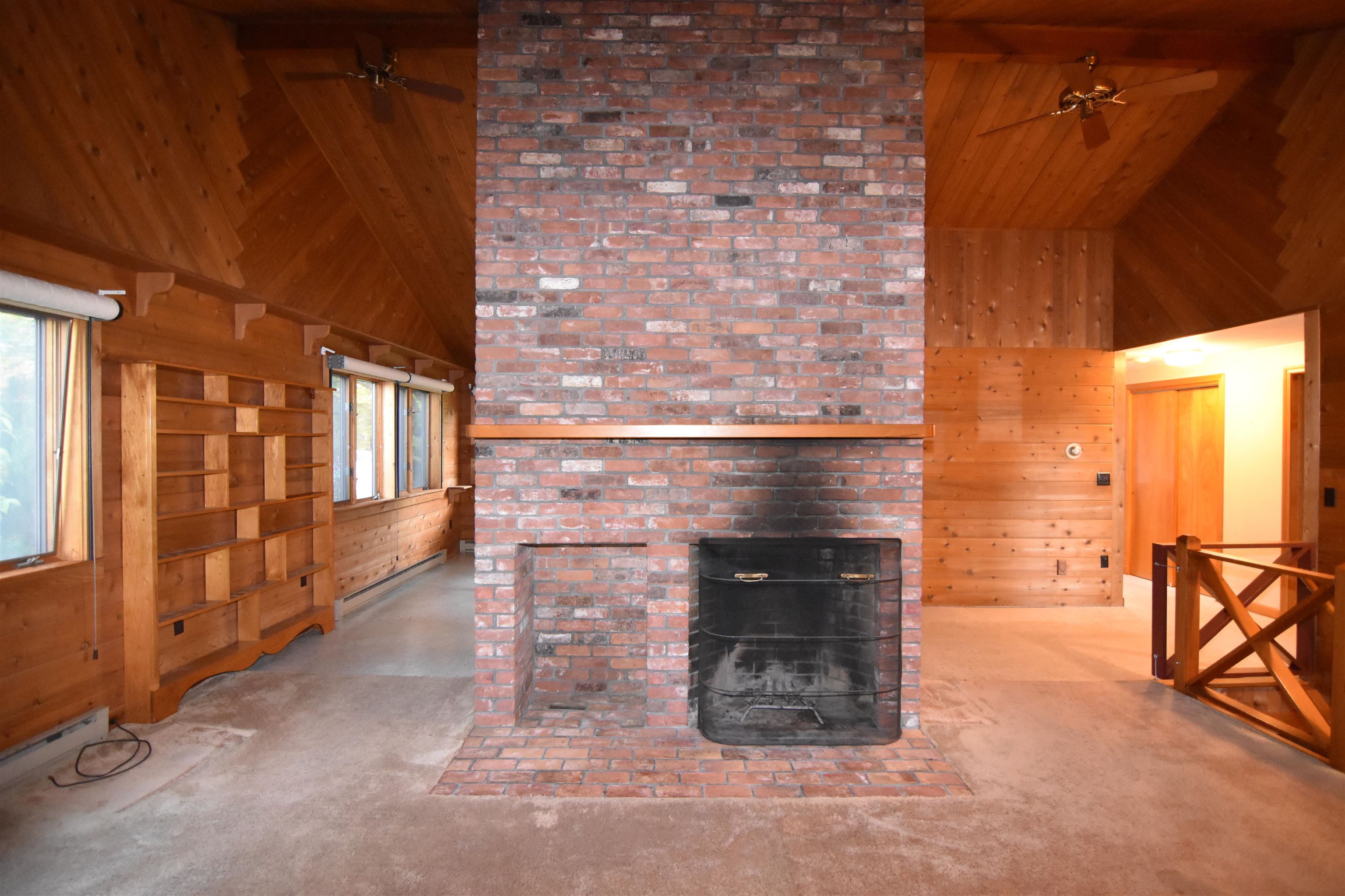 Vacant living room with a tall brick fireplace, vaulted wood ceiling, and built-in shelving.