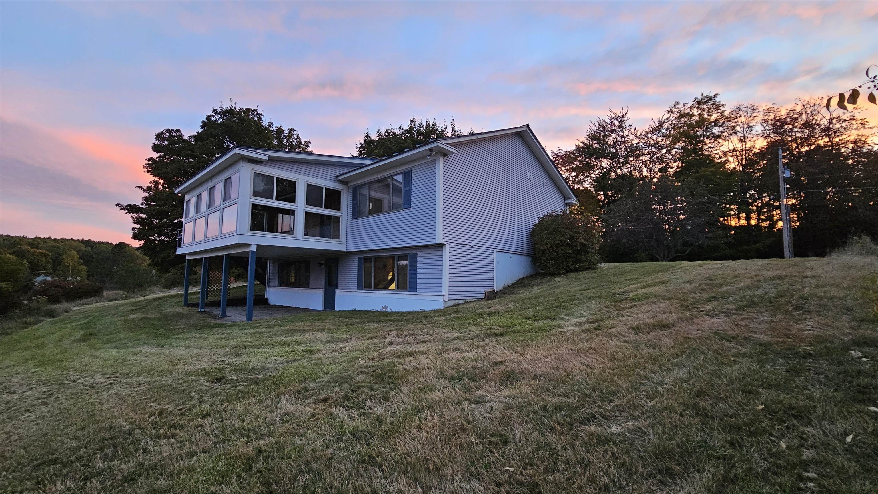 White two-story house with a large windowed section on a sloping lawn beneath a colorful dusk sky.
