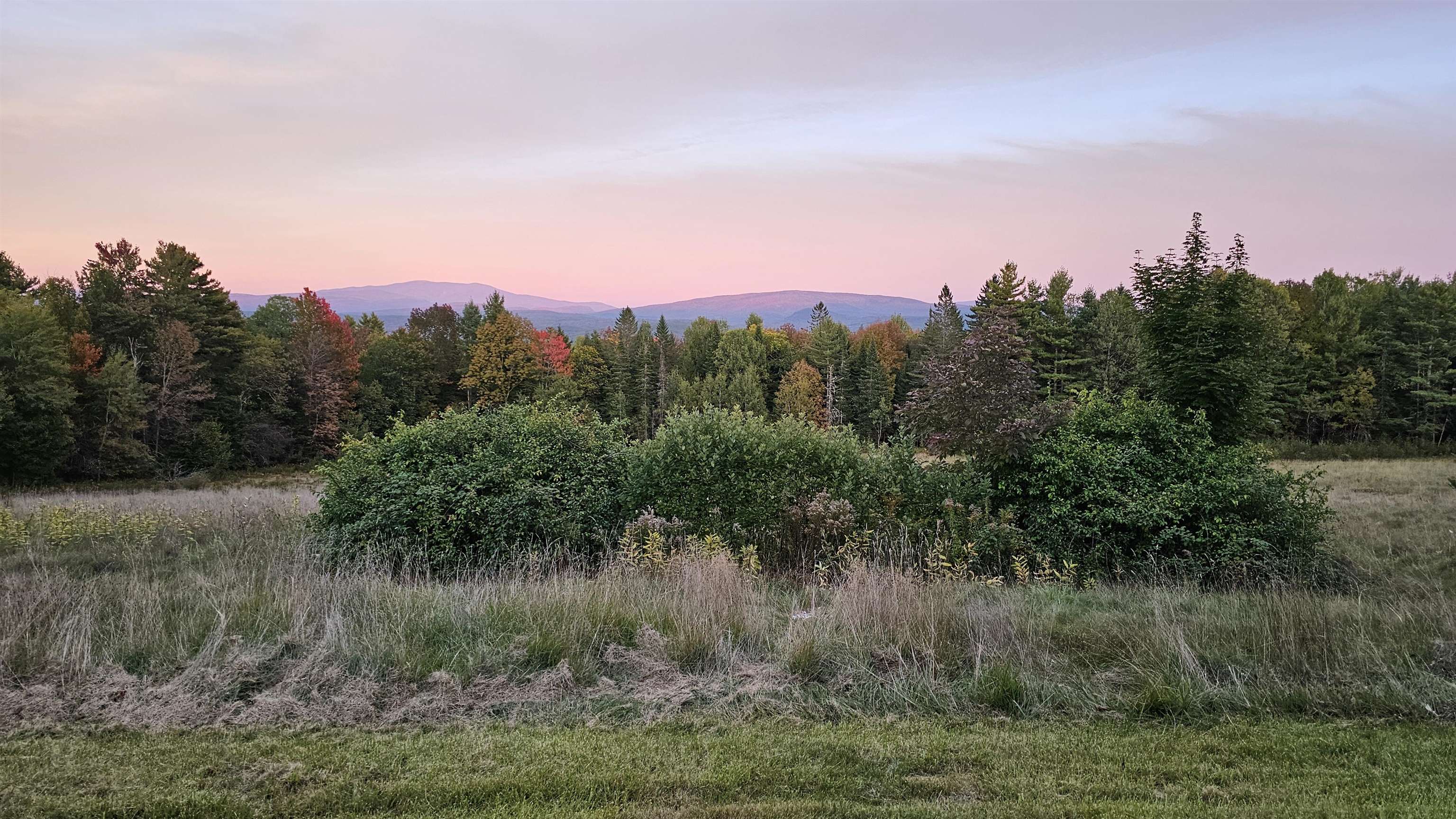 Open field and autumn trees with distant mountain ridges beneath a pink evening sky.