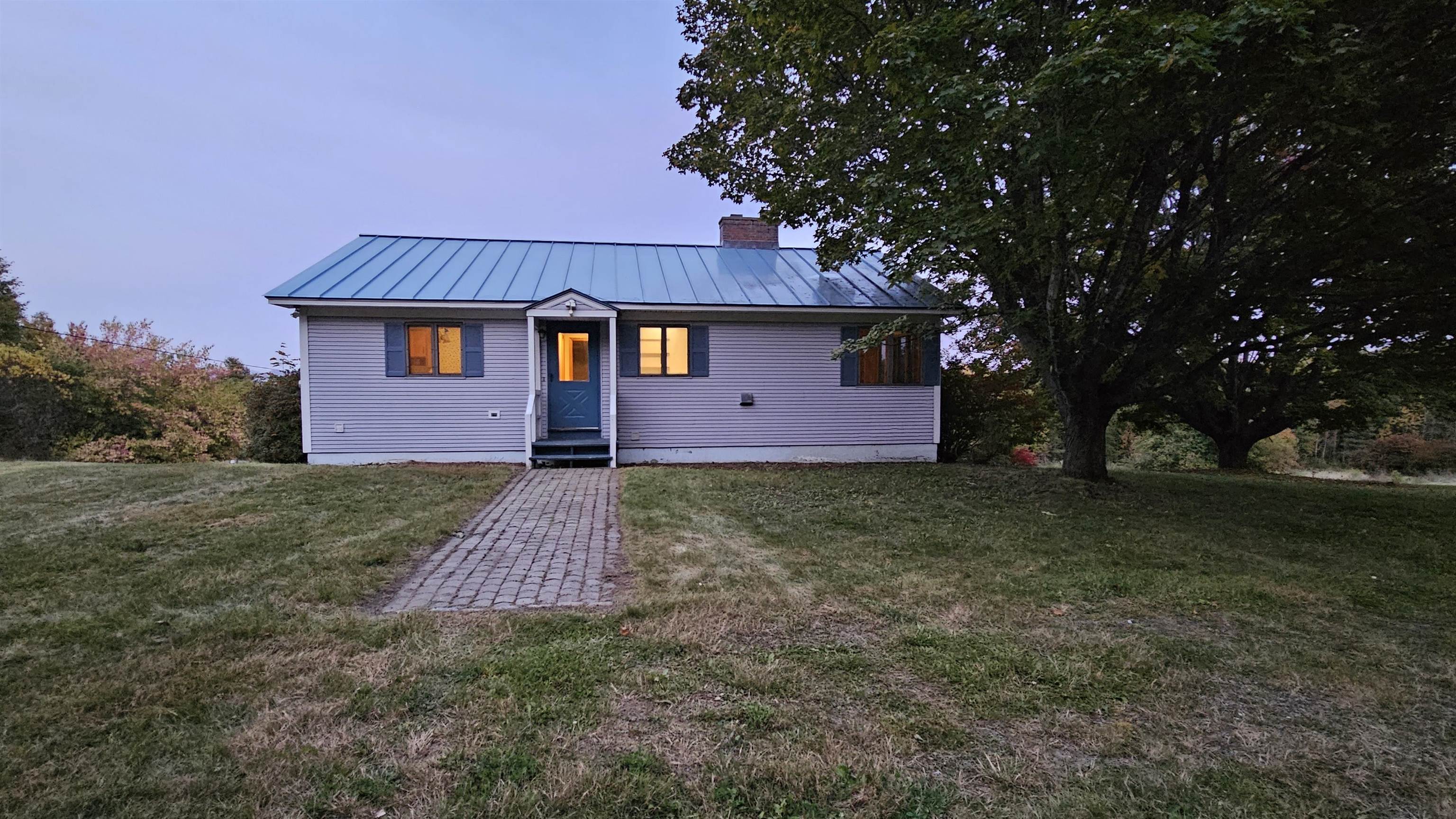Single-story gray house with a blue metal roof, lit windows, and a brick walkway at dusk.