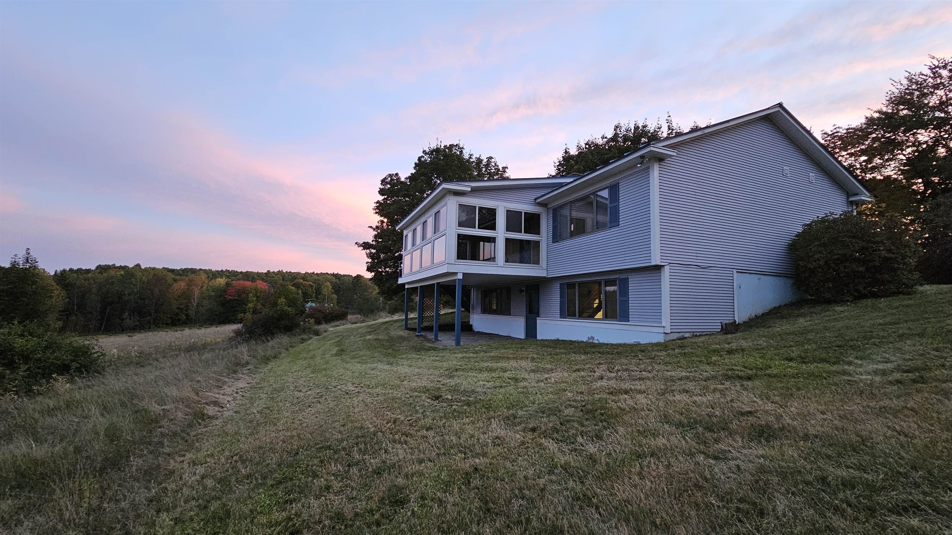 White two-story house with a projecting sunroom on a grassy hillside at dusk.