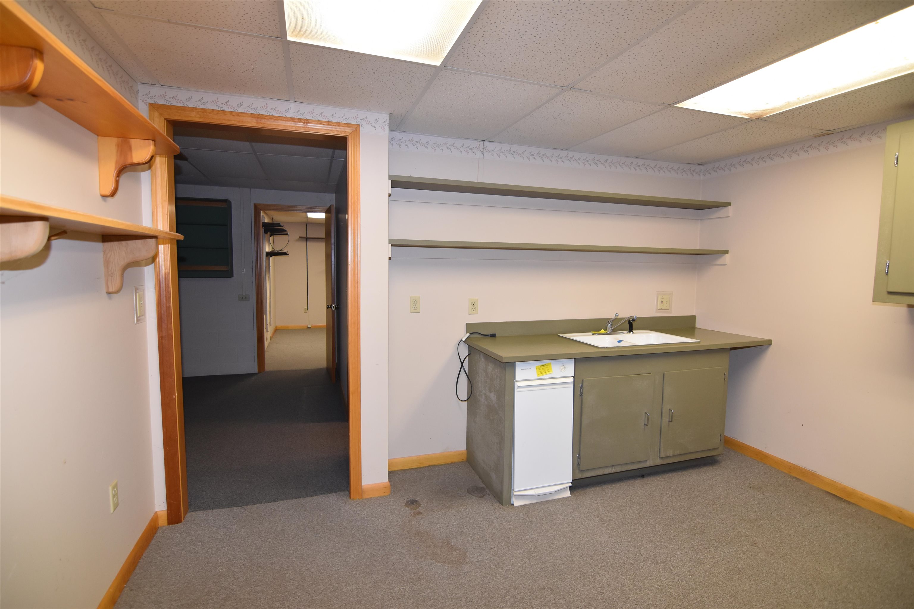 Empty kitchenette with a sink, olive-green cabinets, wall shelves, and doorways to adjoining rooms.