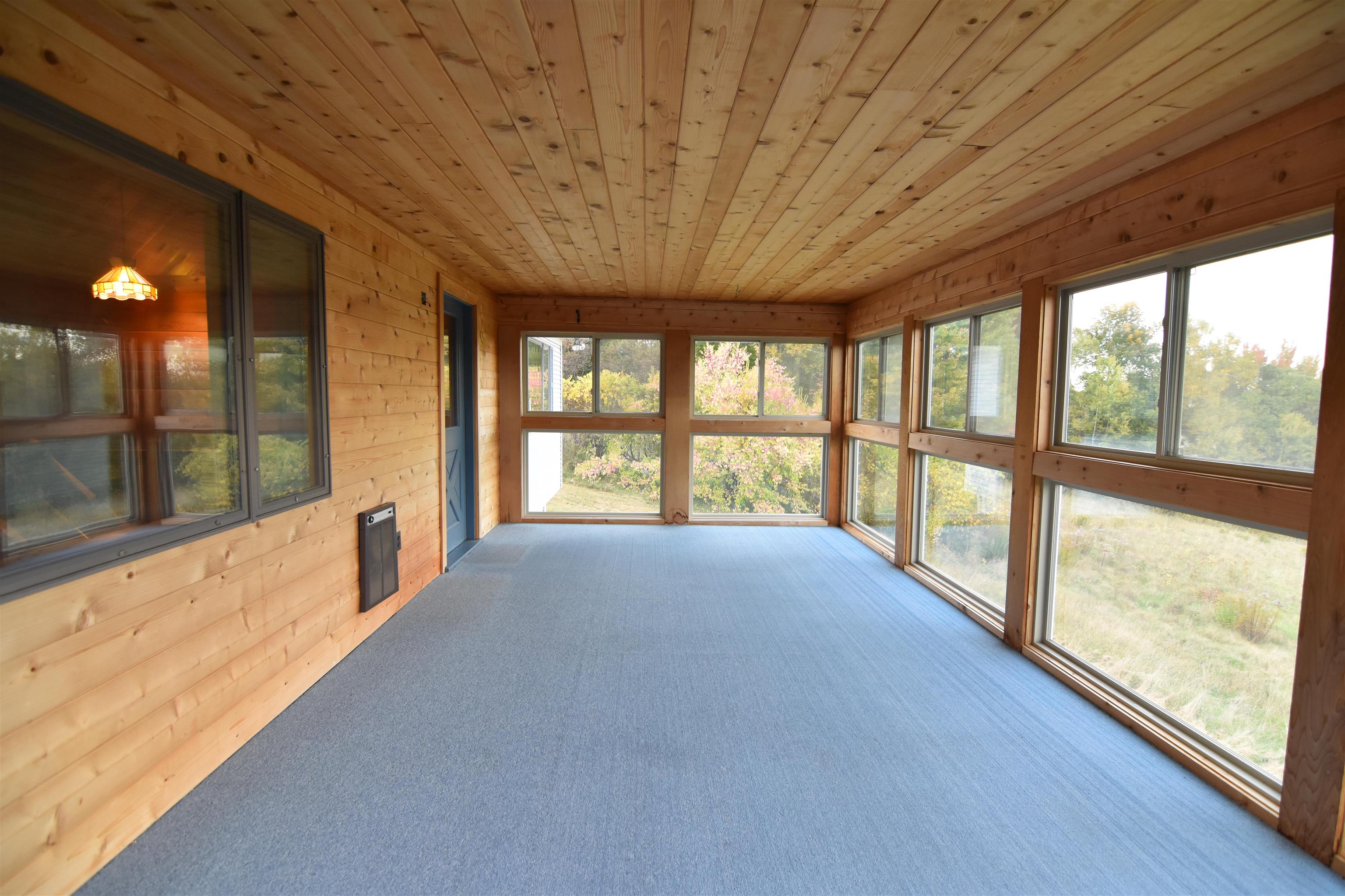 Vacant wood-paneled sunroom with blue-gray carpet and large windows overlooking autumn foliage.