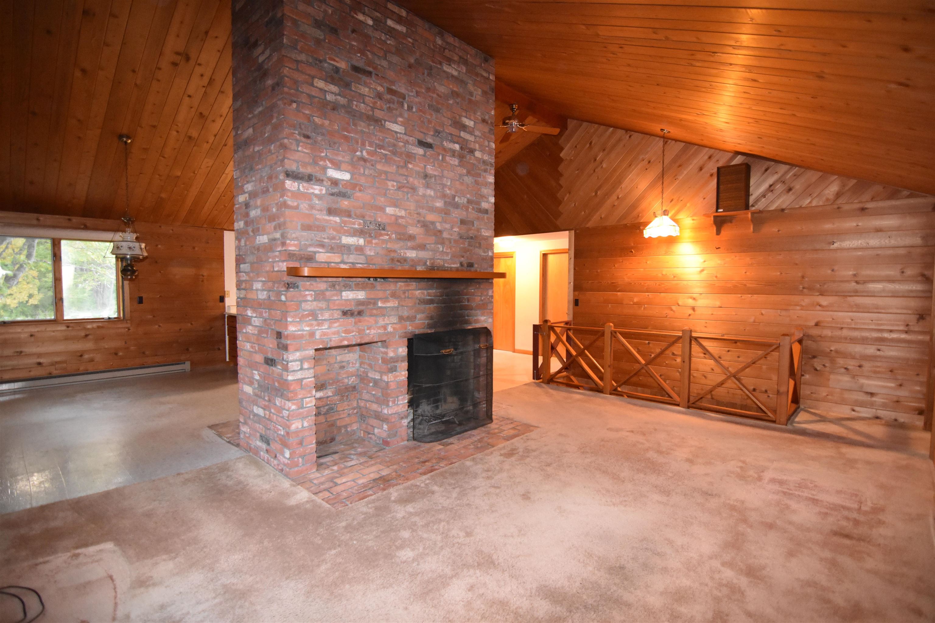 Living room with a tall brick fireplace, vaulted wood ceiling, and railing around a stair opening.
