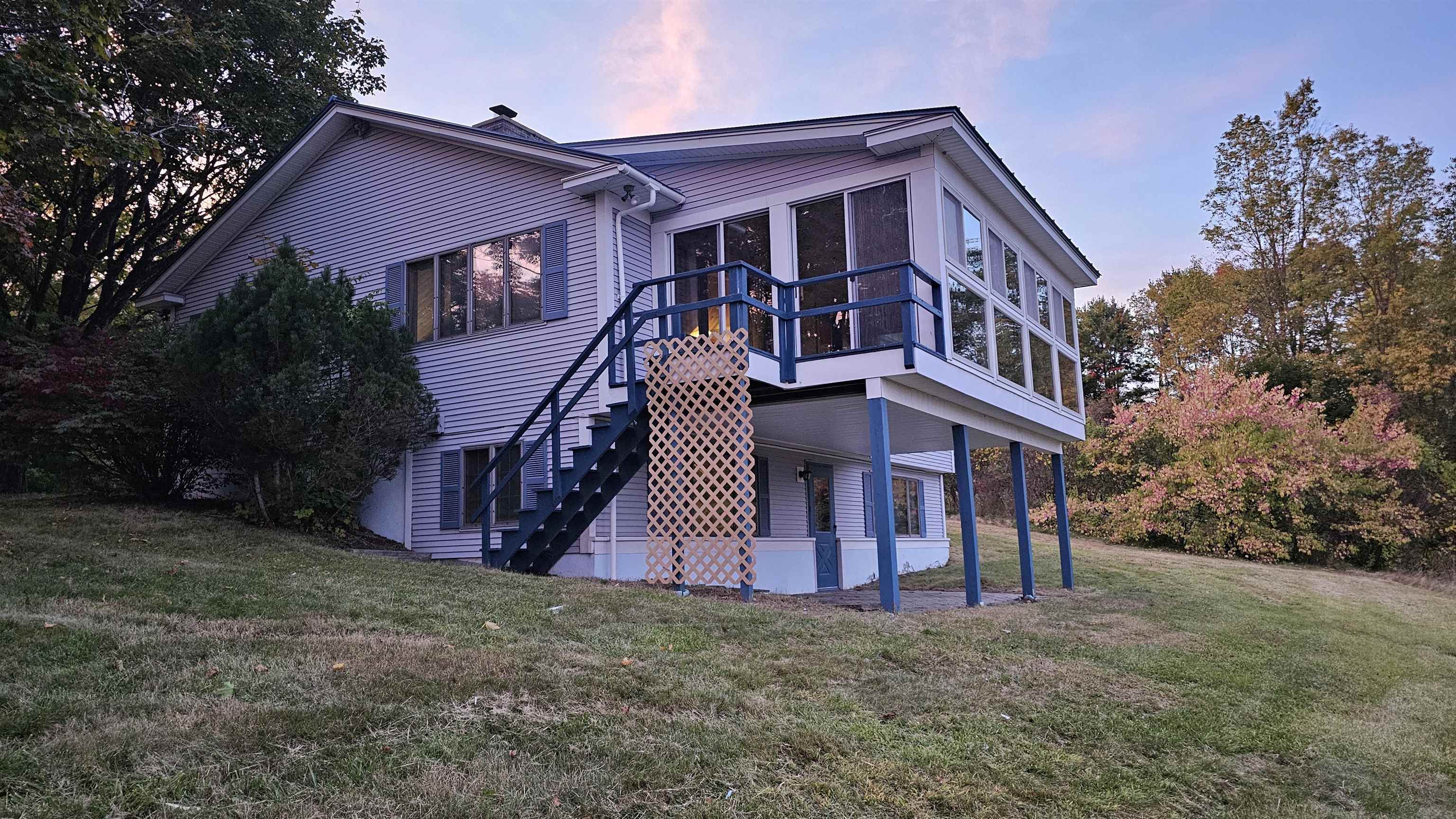House with a raised glass sunroom, blue railings, and exterior stairs amid autumn trees.