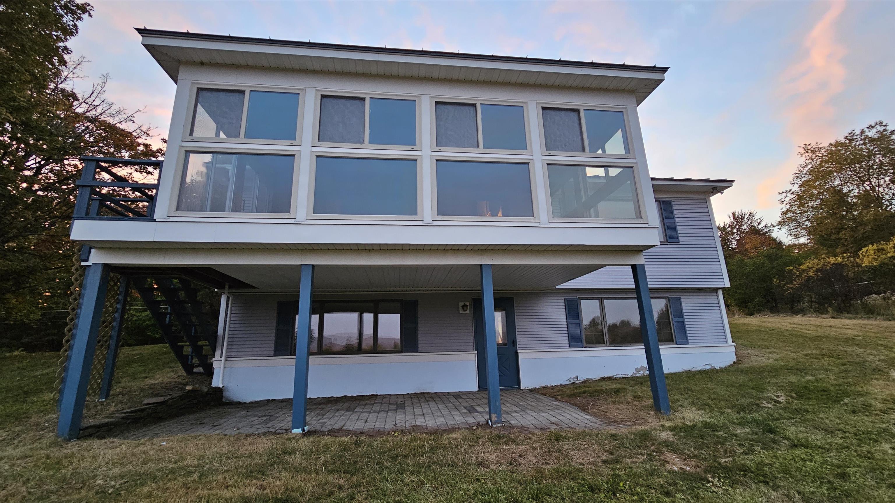 Raised gray house with a wide, glass-fronted sunroom and blue support posts at dusk.