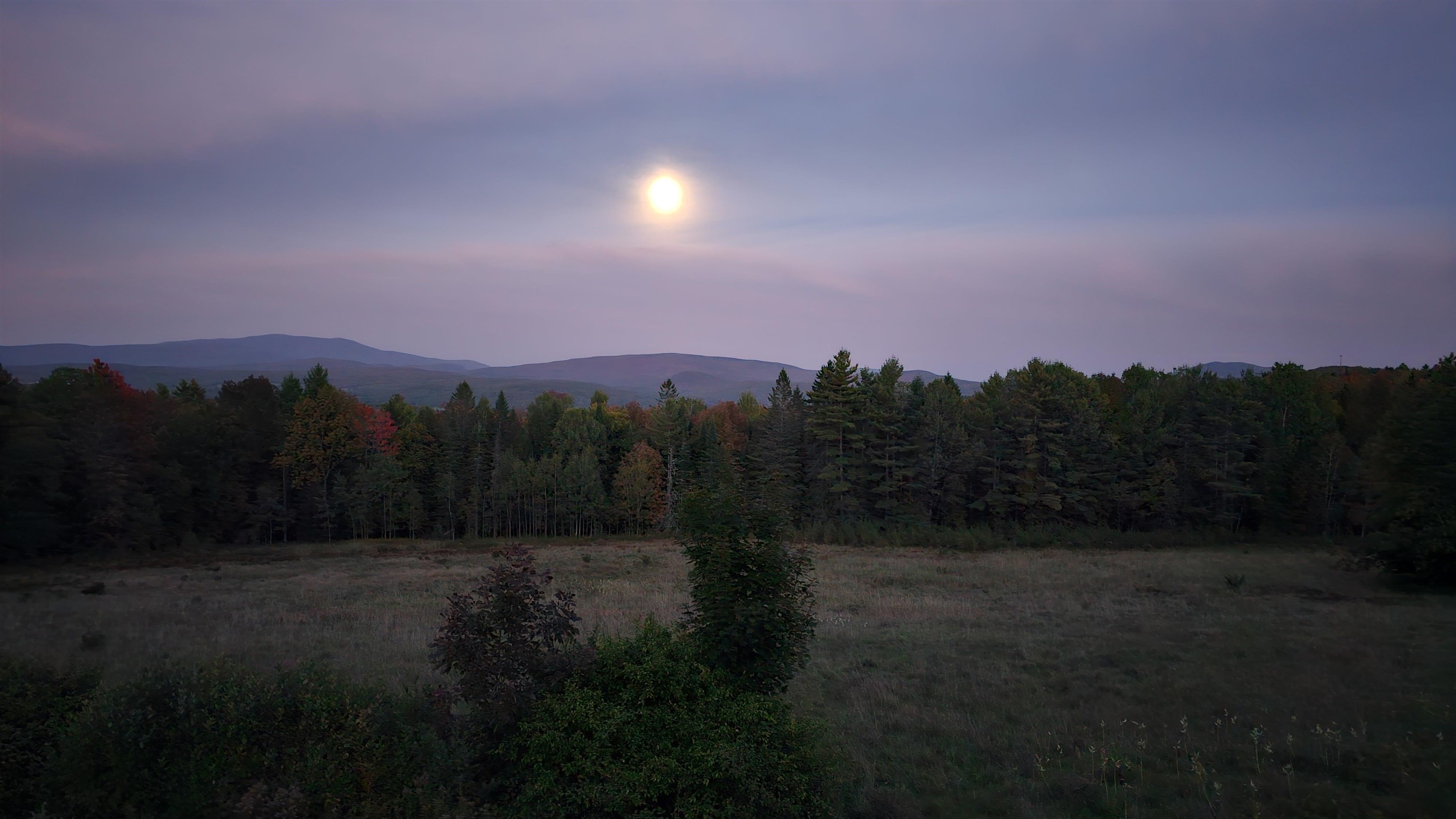 Open meadow and autumn forest with distant mountain ridges beneath the setting sun.
