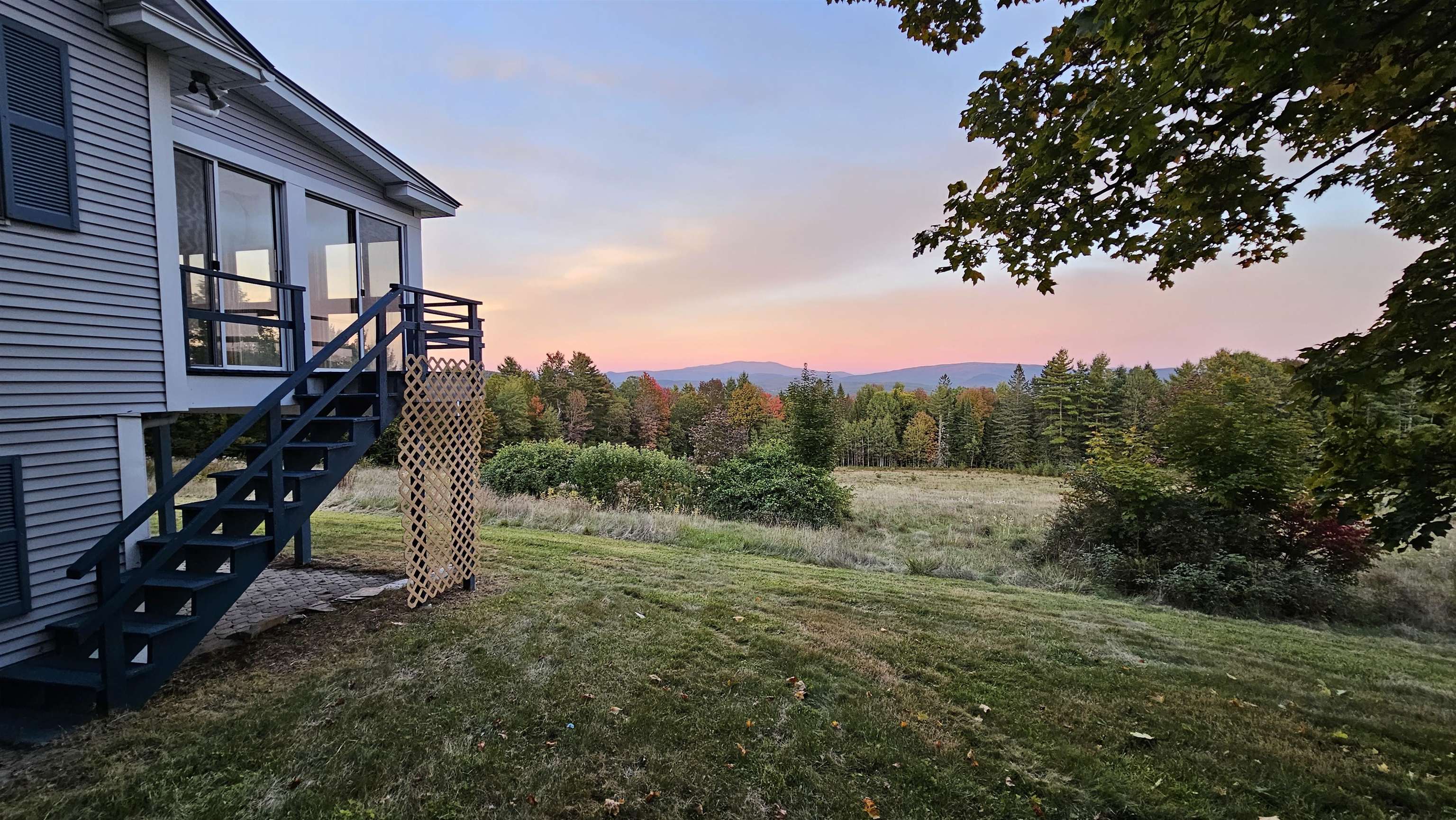 Gray-sided home and deck overlook an autumn meadow and distant mountains at sunset.