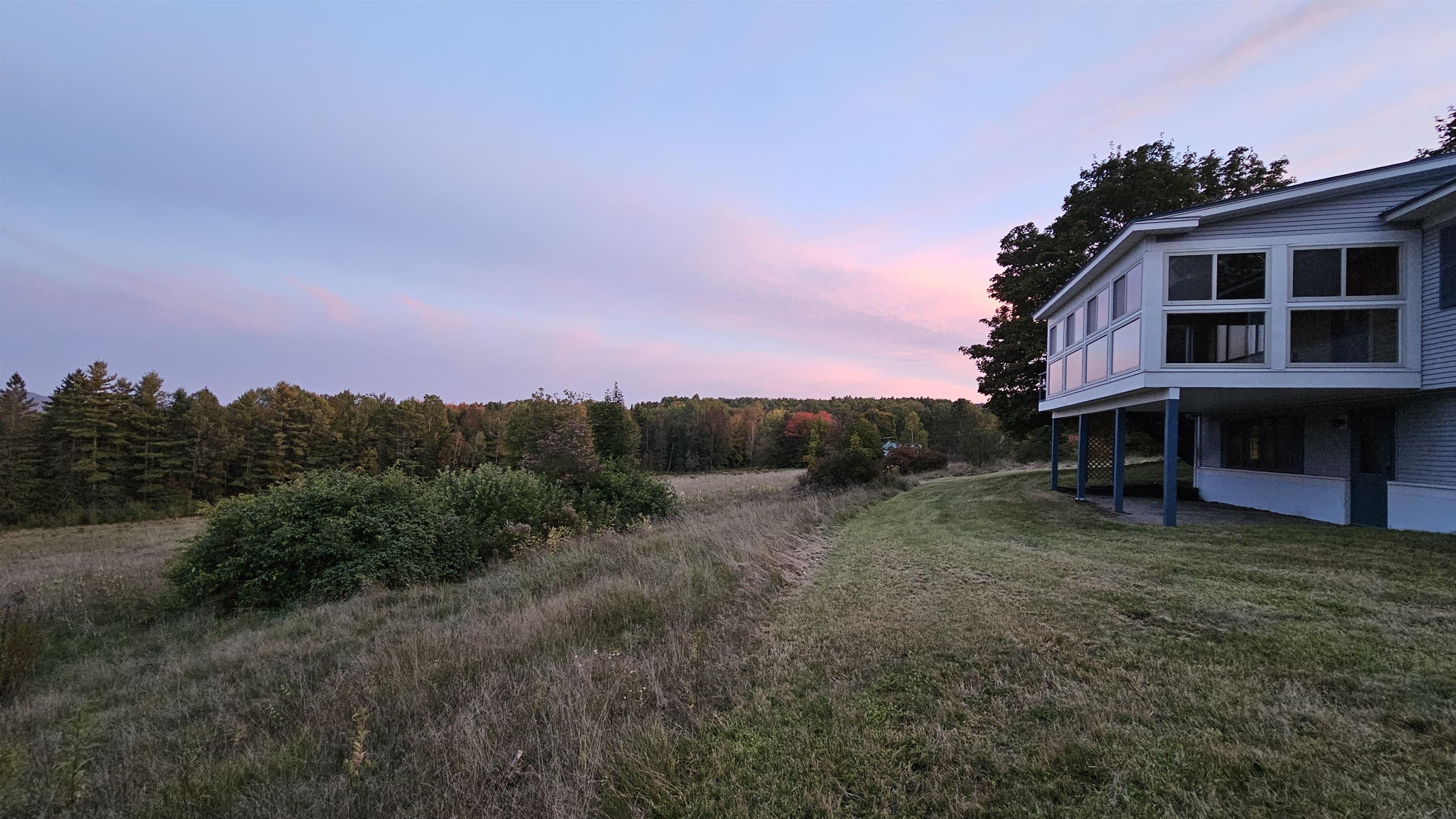 Raised house with a glass-enclosed sunroom beside a grassy field at pink dusk.