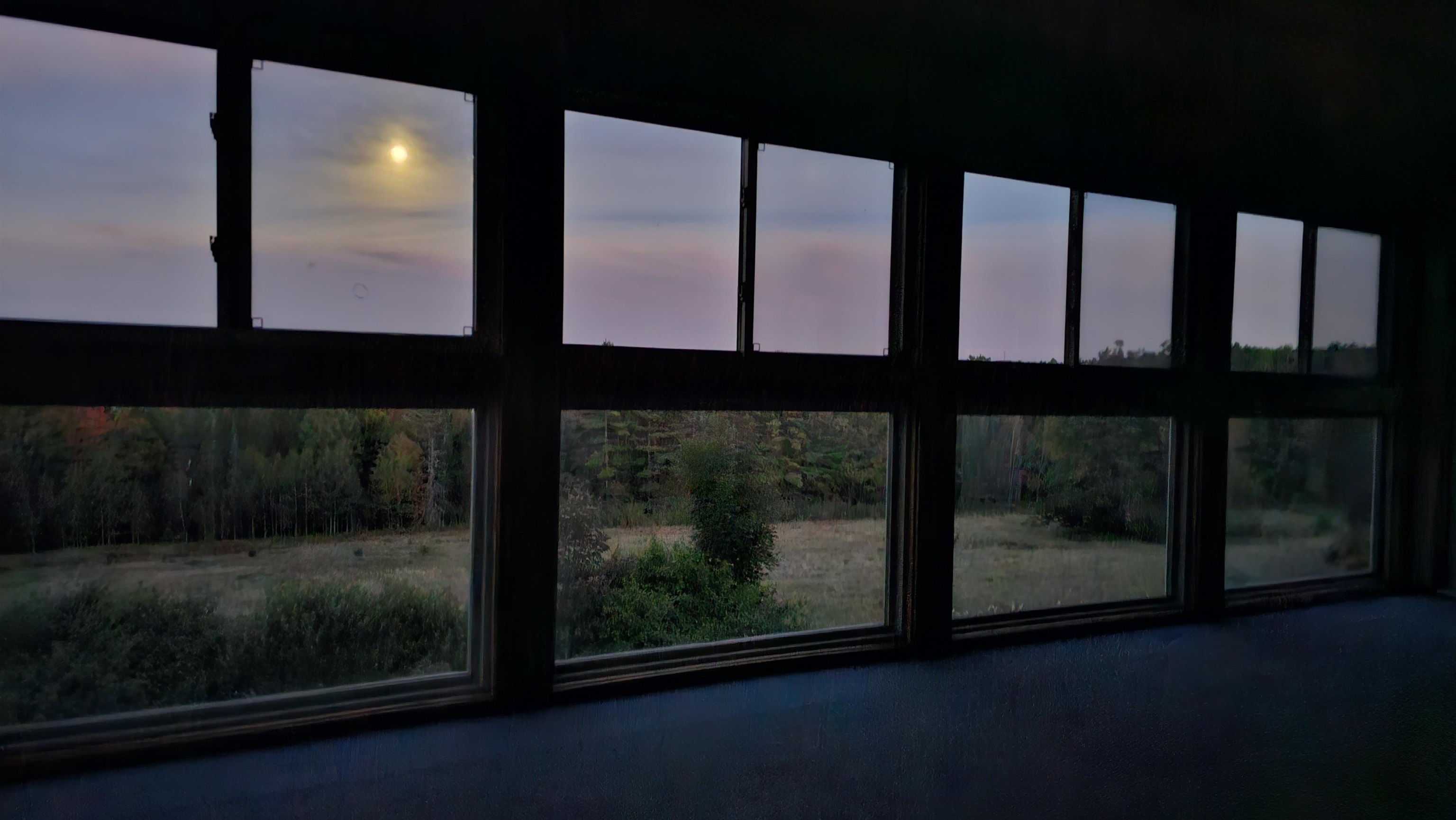 Wide interior windows overlook a grassy field and autumn woods beneath a pink dusk sky.