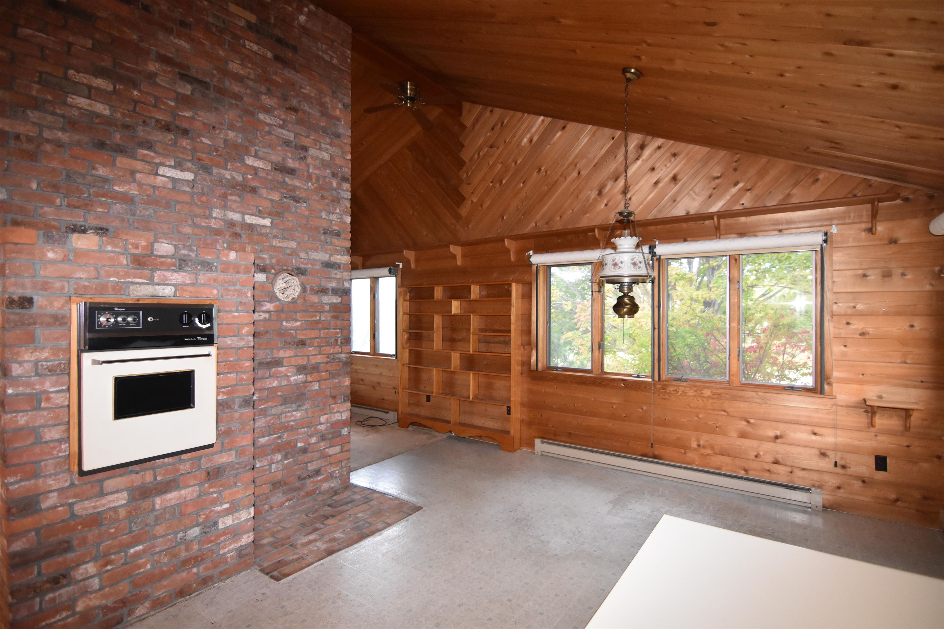 Wood-paneled room with vaulted ceiling, brick fireplace and oven, built-in shelves, and broad windows.