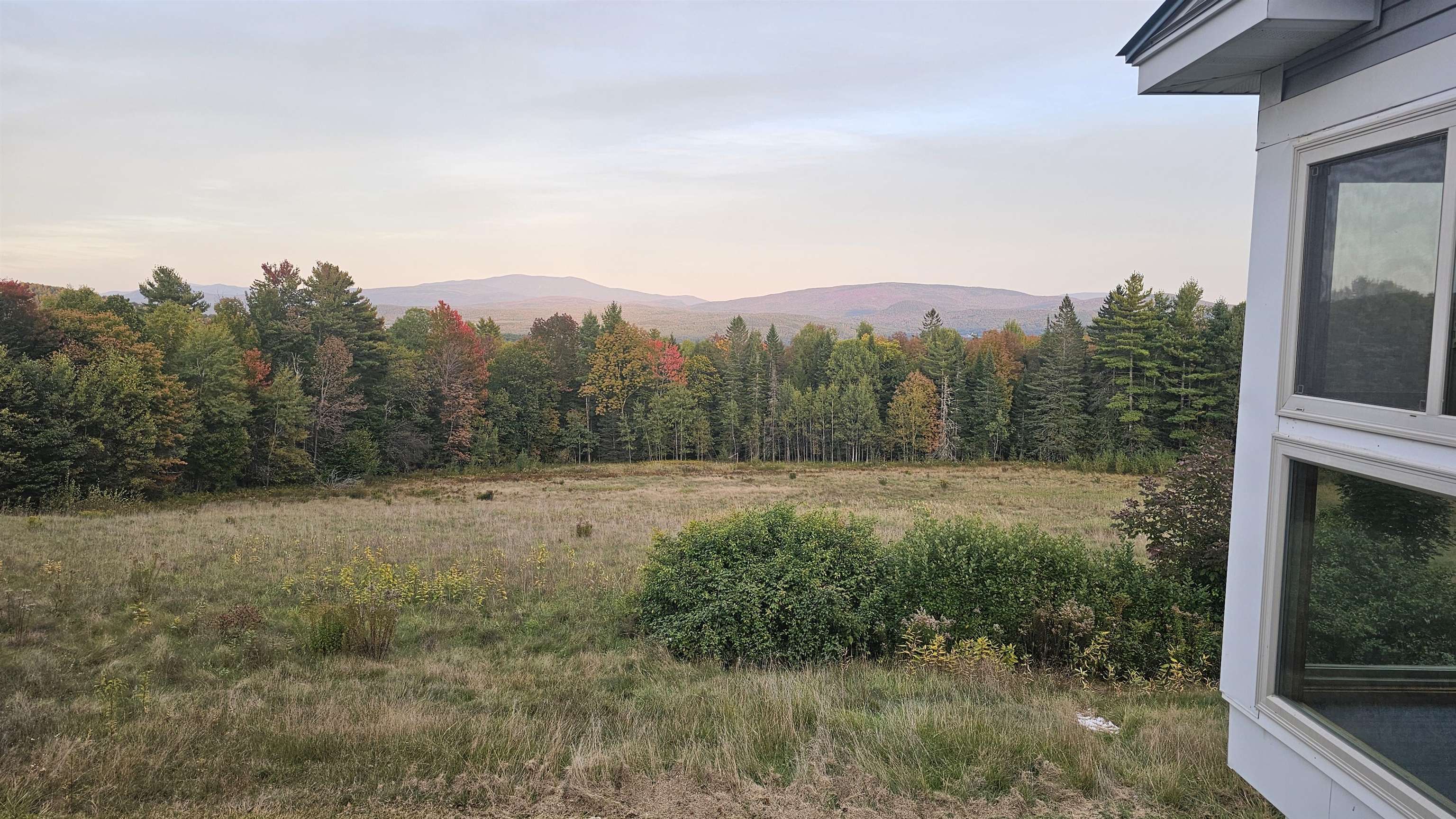Meadow and autumn woodland with mountain ridges beyond, seen beside a white house.