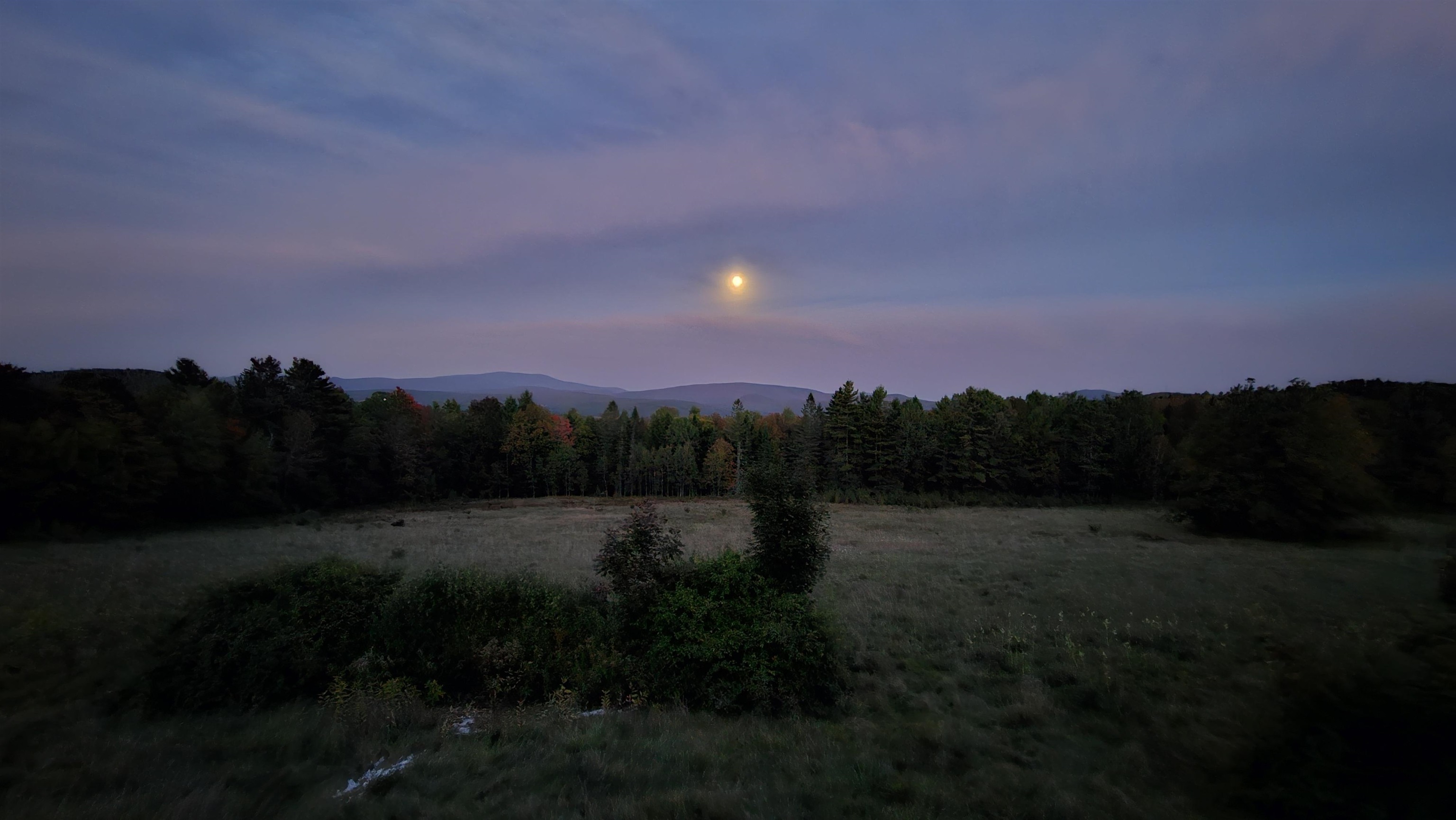 Open field bordered by trees, with distant mountain ridges beneath a glowing moon at dusk.