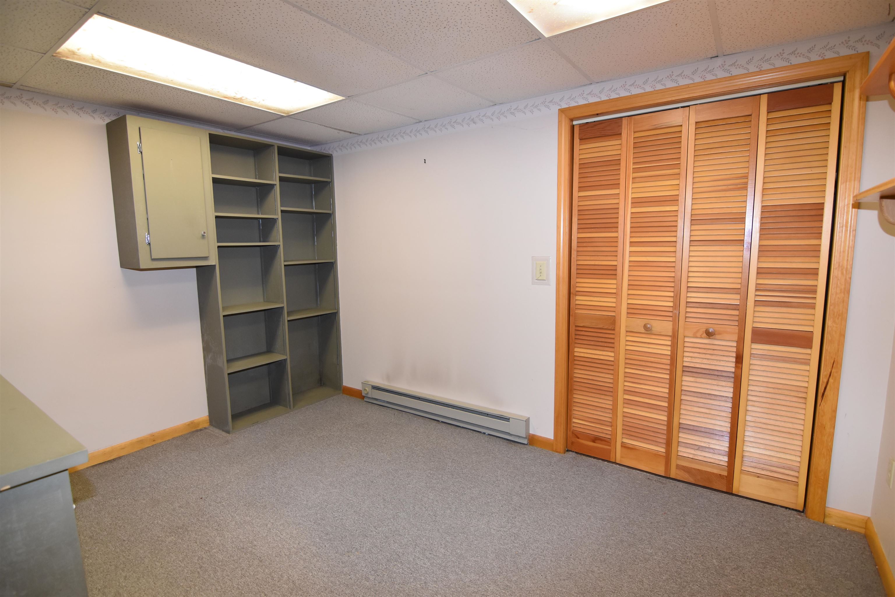 Carpeted room with built-in shelving, louvered closet doors, and a baseboard heater.