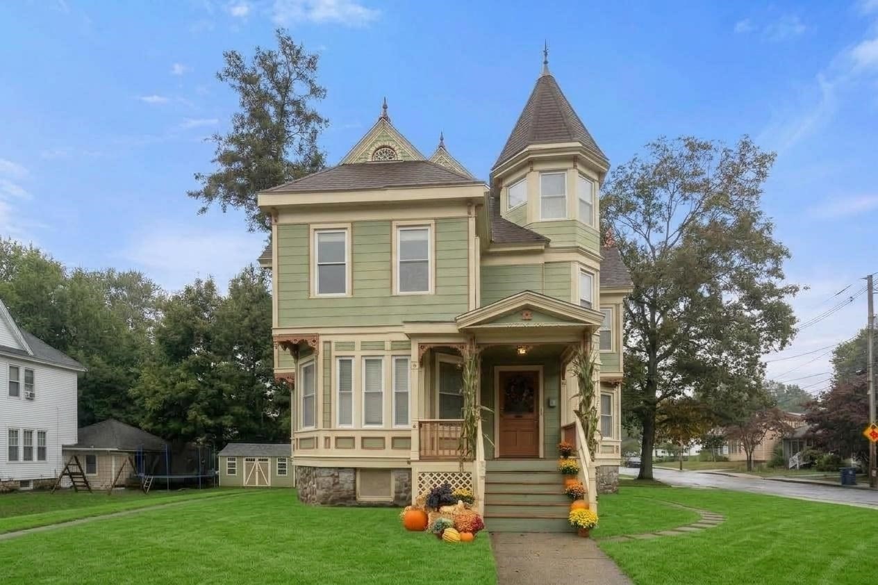 Green Victorian house with a turret, covered porch, and pumpkins by the front steps.