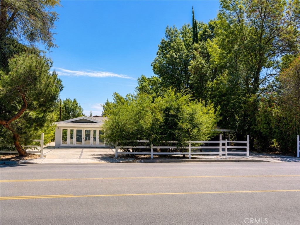 Low modern home behind a white fence, partly screened by mature trees along a sunny street.