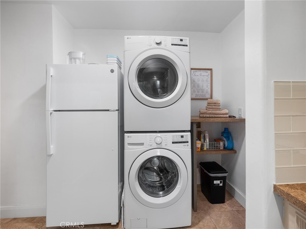 Laundry area with a refrigerator beside stacked front-loading washer and dryer, open shelves, and a black bin.