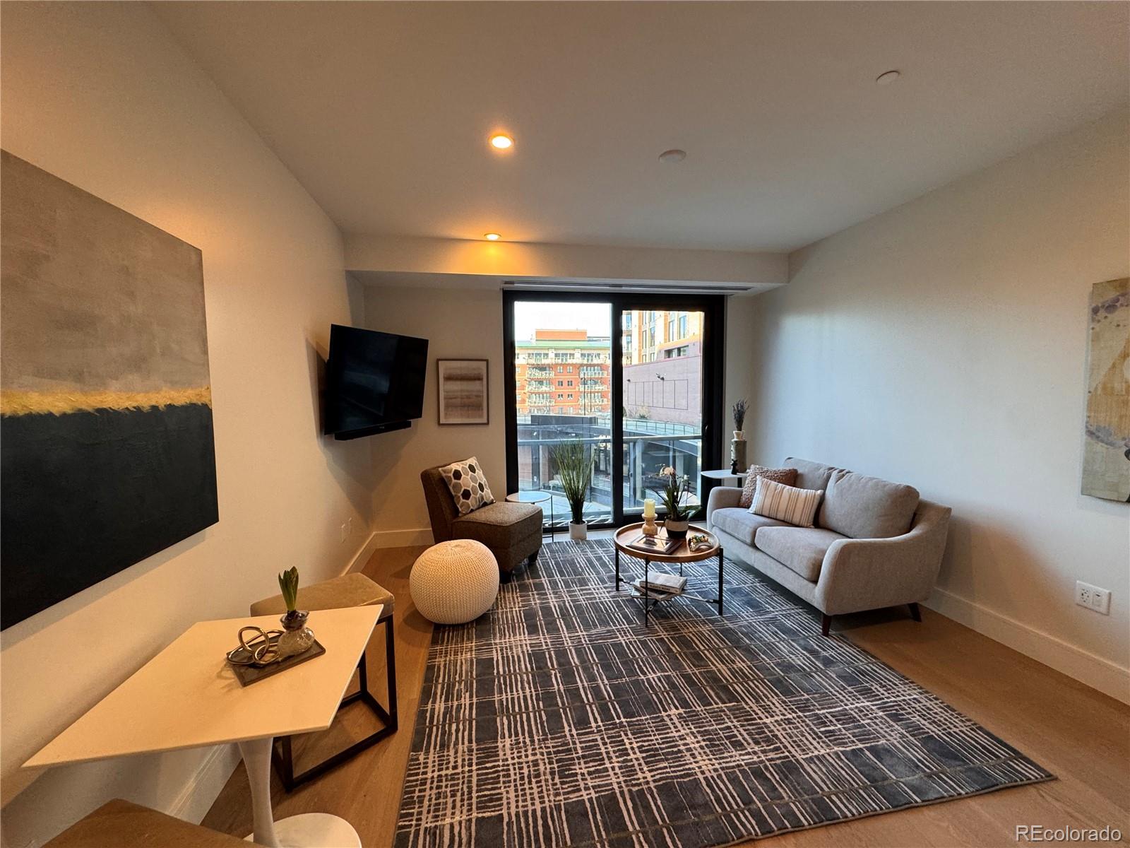 Furnished condo living room with a gray sofa and balcony doors overlooking nearby buildings.