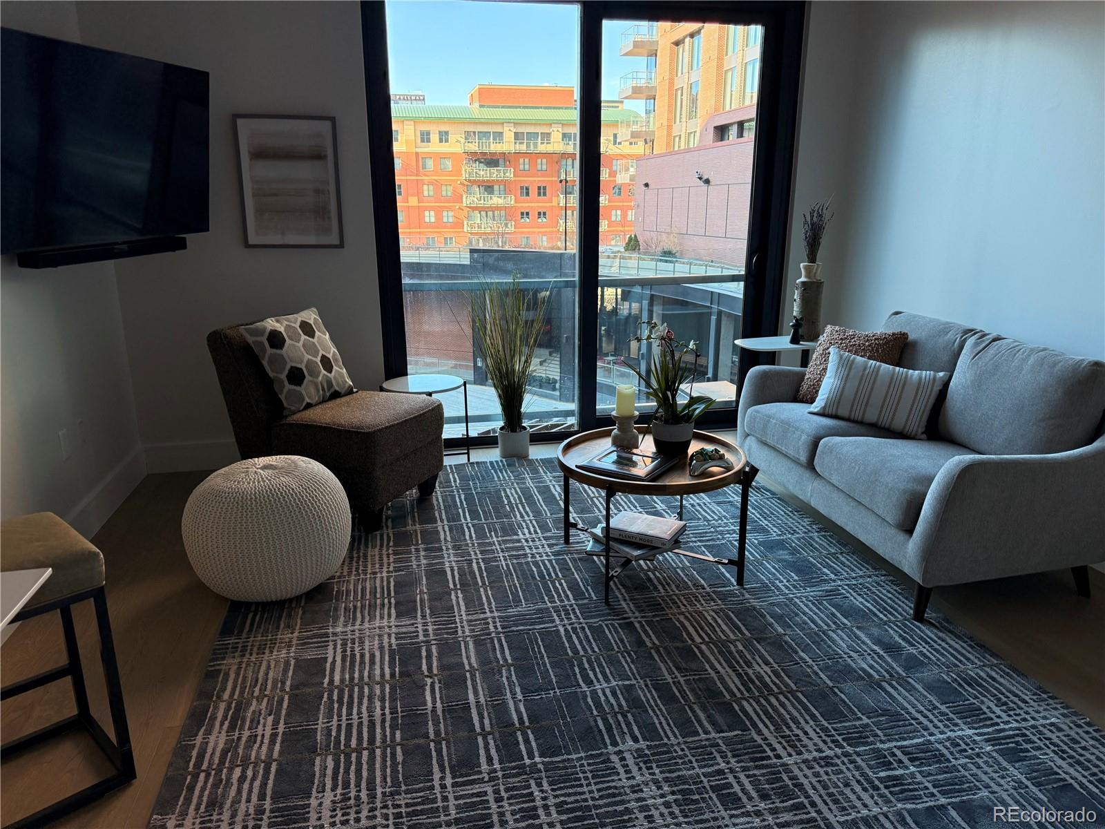 Staged condo living room with a gray sofa and floor-to-ceiling windows overlooking city buildings.