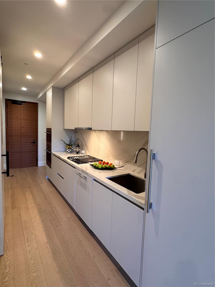 Modern galley kitchen with pale cabinetry, veined stone backsplash, gas cooktop, and dark sink.