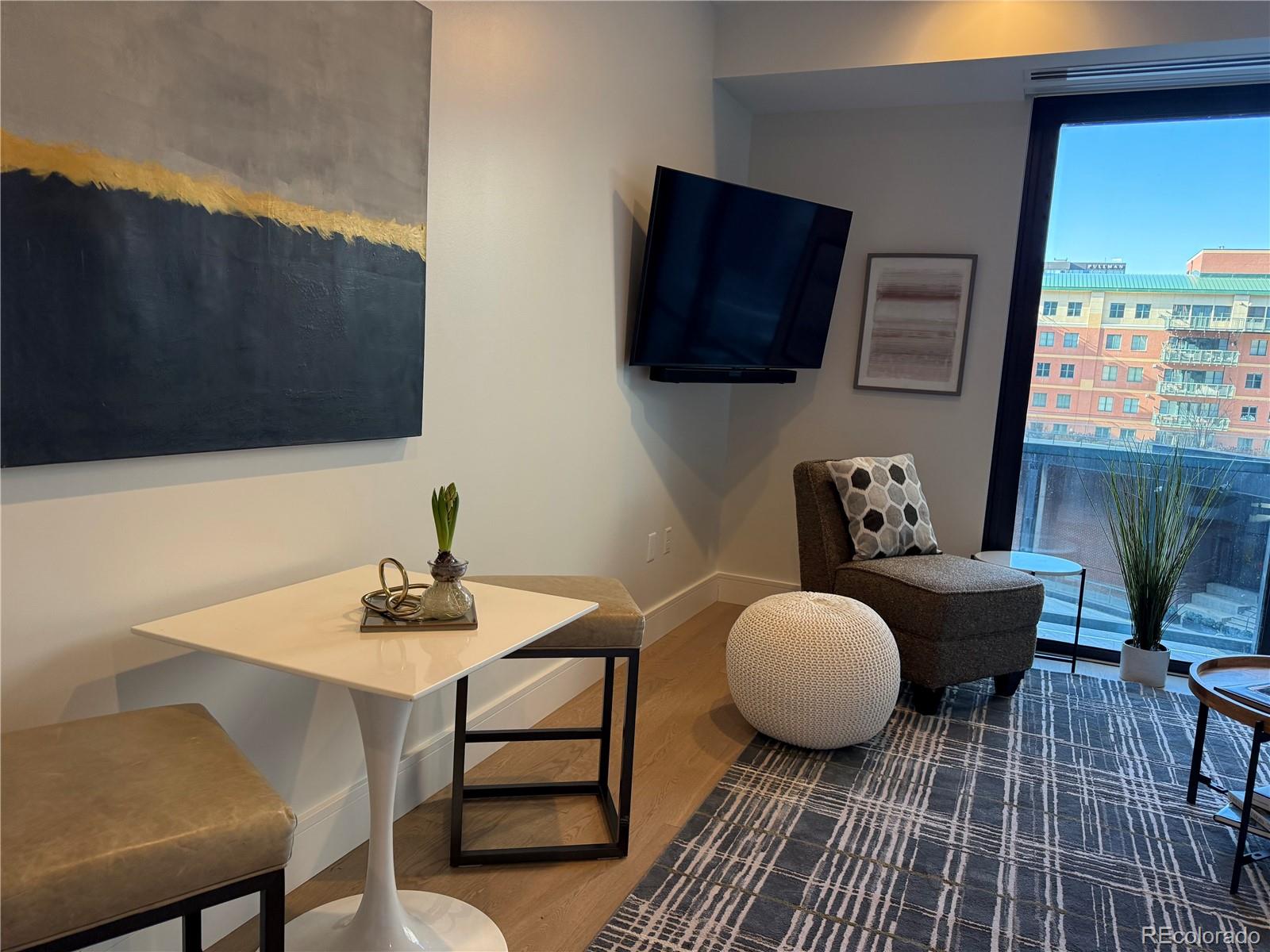 Staged condo living area with a white tulip table, lounge chair, and large windows overlooking nearby buildings.