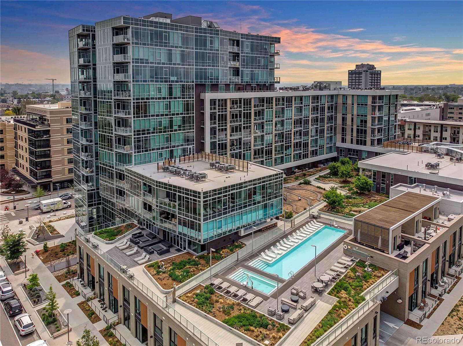 Modern glass condominium complex with balconies, landscaped rooftop terraces, and a swimming pool at sunset.