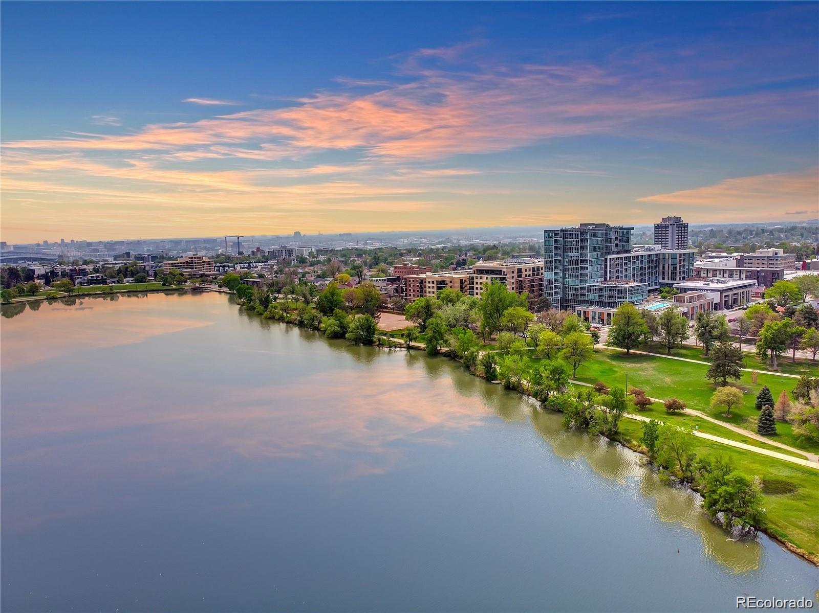 Aerial lake view toward Denver high-rises, with a green shoreline park under a pink-and-gold sunset sky.