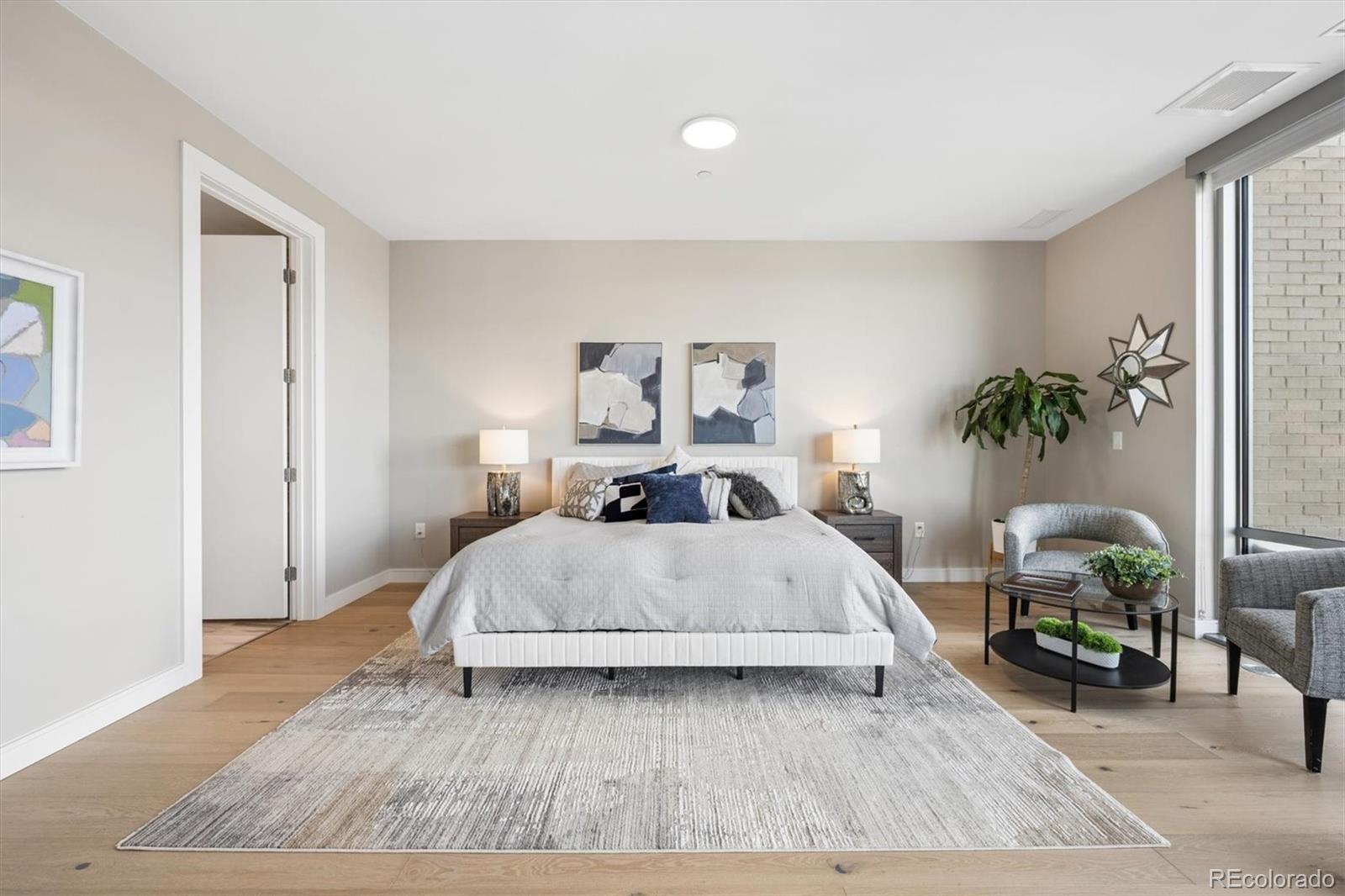 Staged bedroom with a large bed, light wood floors, and a bright sitting area beside a floor-to-ceiling window.