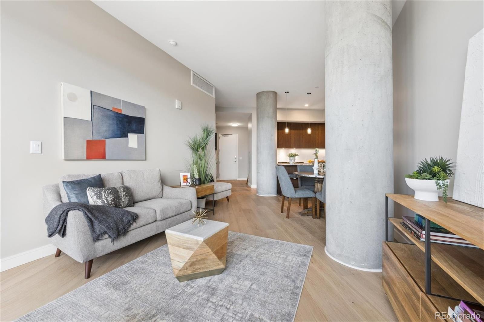 Staged condo living room with a gray sofa, exposed concrete columns, and dining area beyond.