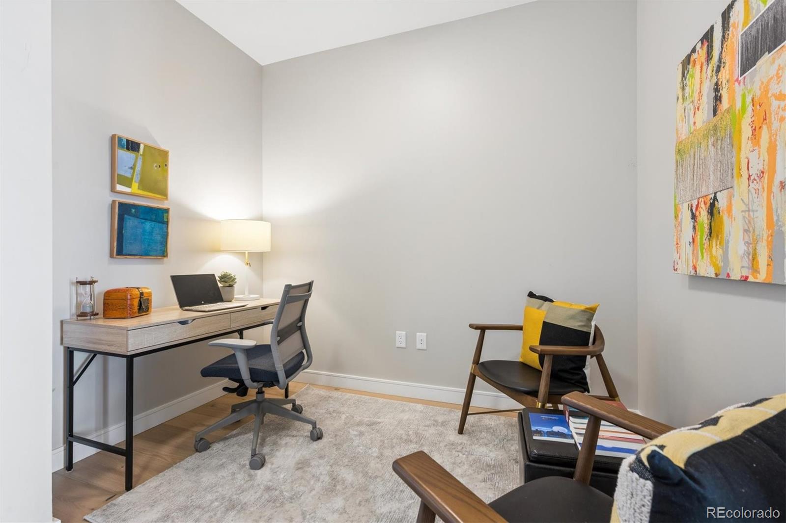 Furnished home office with a desk and laptop, colorful wall art, and wood-framed lounge chairs.