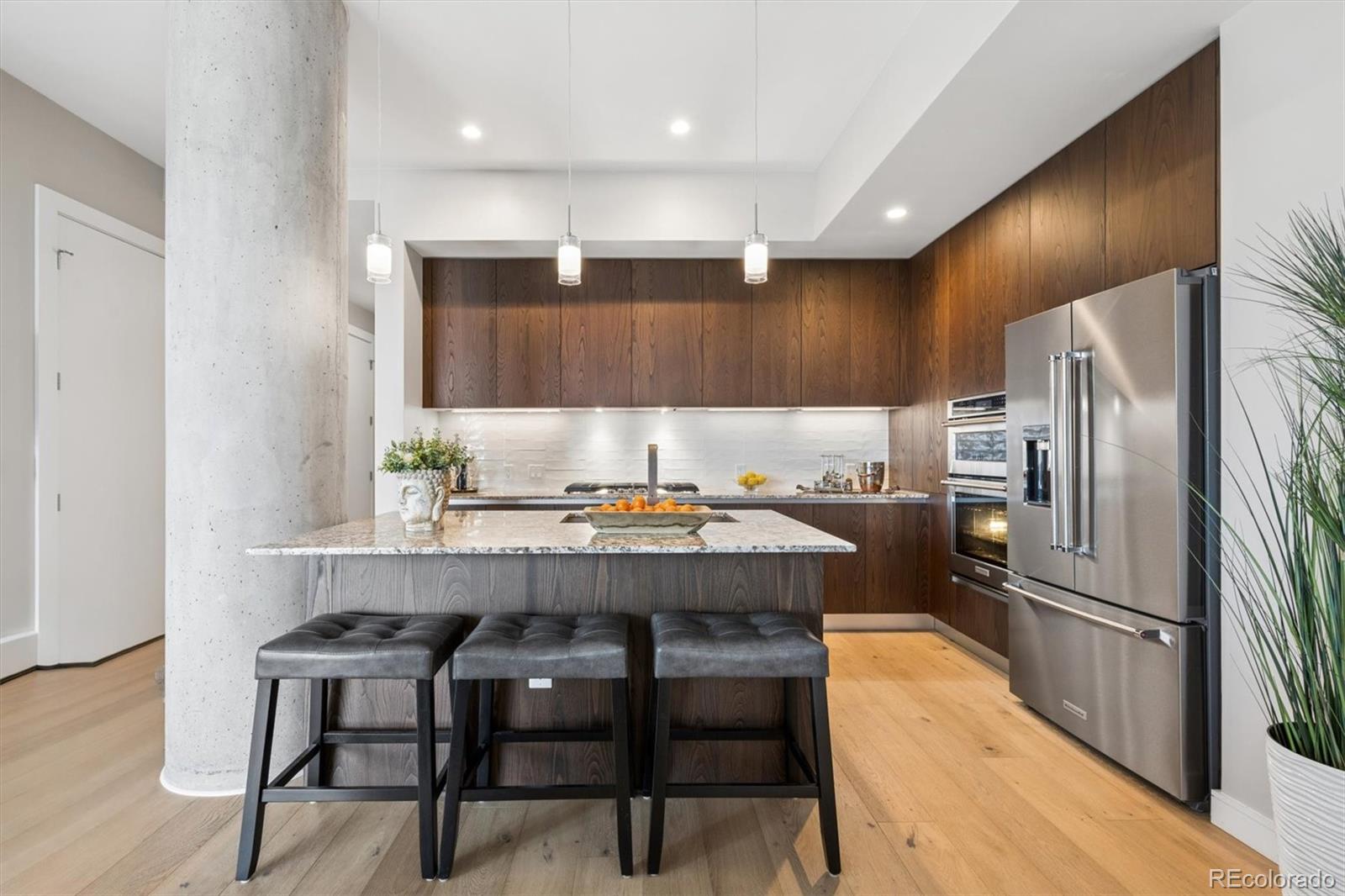Modern condo kitchen with a stone island, dark wood cabinets, three gray stools, and stainless steel appliances.