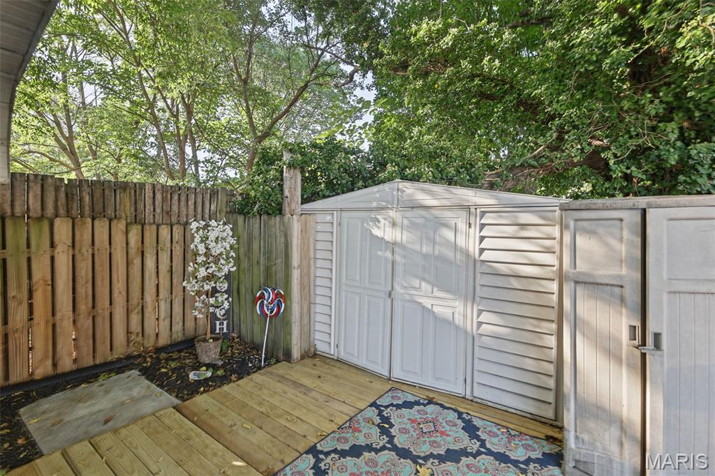 Backyard deck with a white storage shed, colorful rug, wooden fencing, and mature trees.