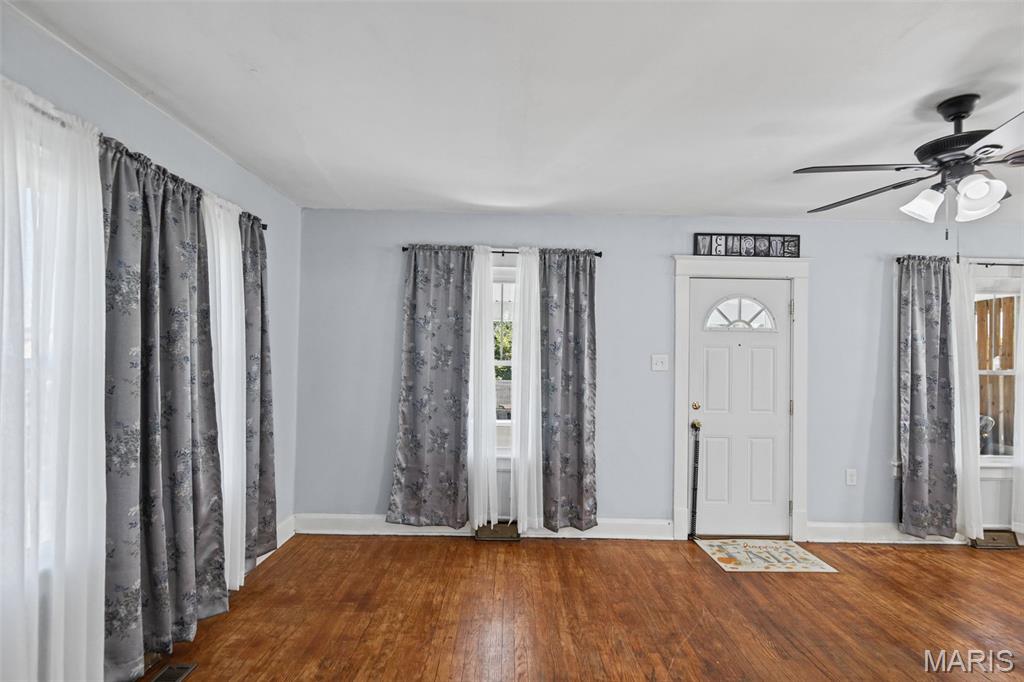 Unfurnished interior with polished wood floors, patterned curtains, a white entry door, and ceiling fan.