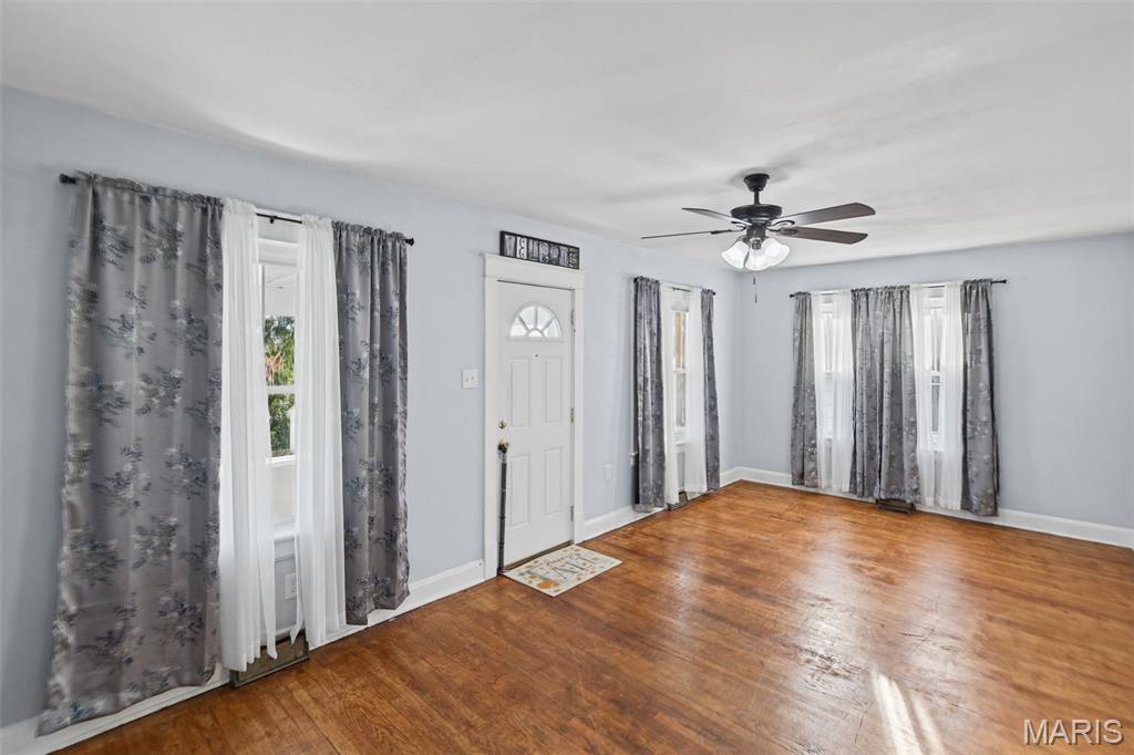 Unfurnished room with hardwood floors, curtained windows, a white front door, and a ceiling fan.