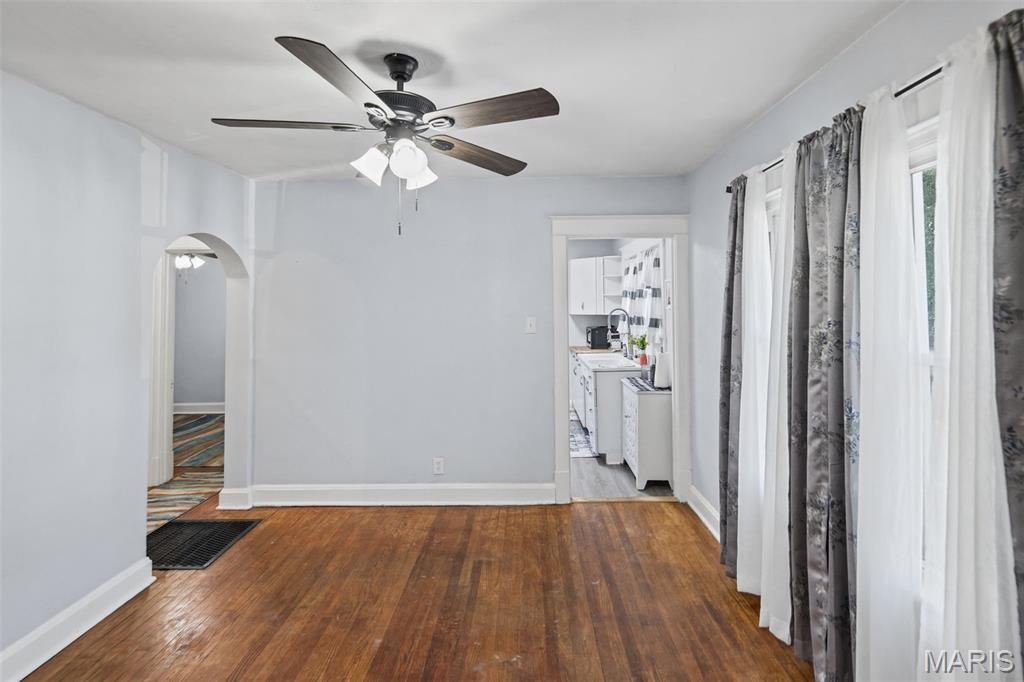 Empty room with hardwood floors, a ceiling fan, and curtained windows beside the kitchen doorway.