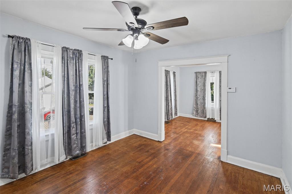 Empty room with hardwood floors, curtained windows, and a ceiling fan, opening into another room.