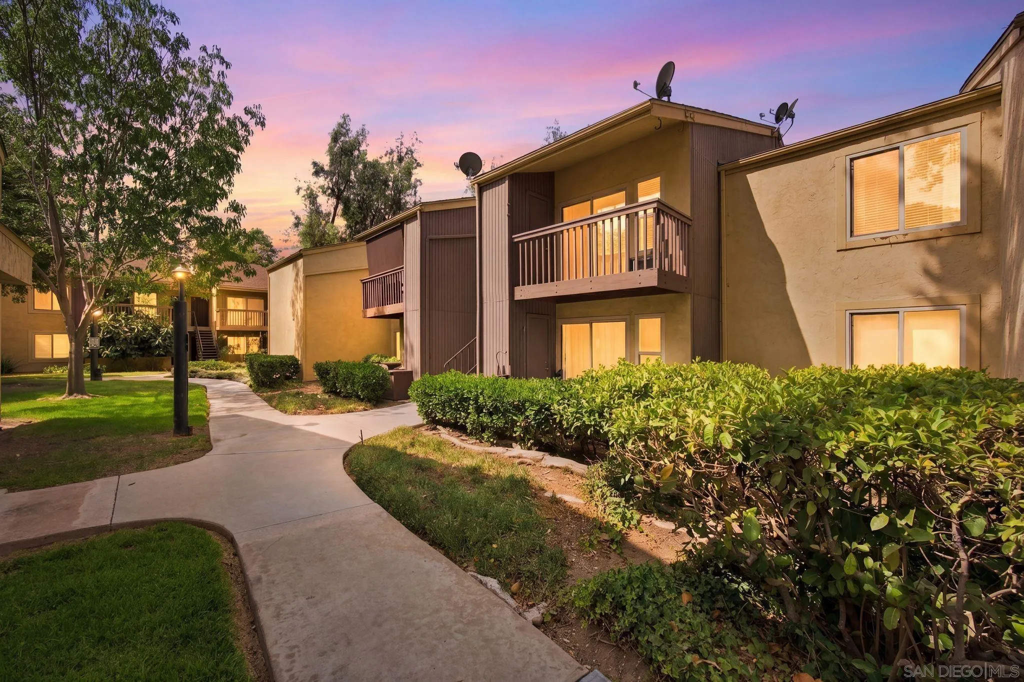 Multi-unit residential building with balconies and landscaped hedges beside a curving walkway at dusk.