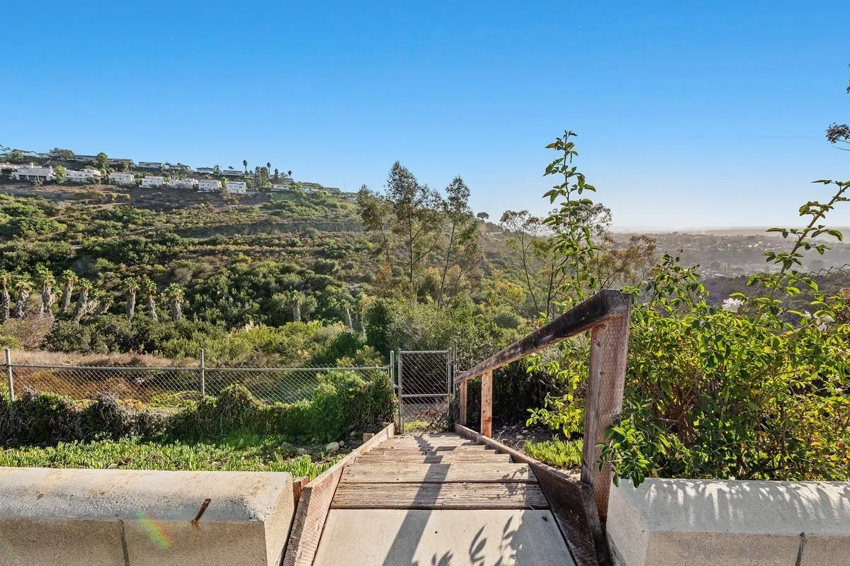 Wooden steps descend toward a fenced, brush-covered hillside with homes in the distance.