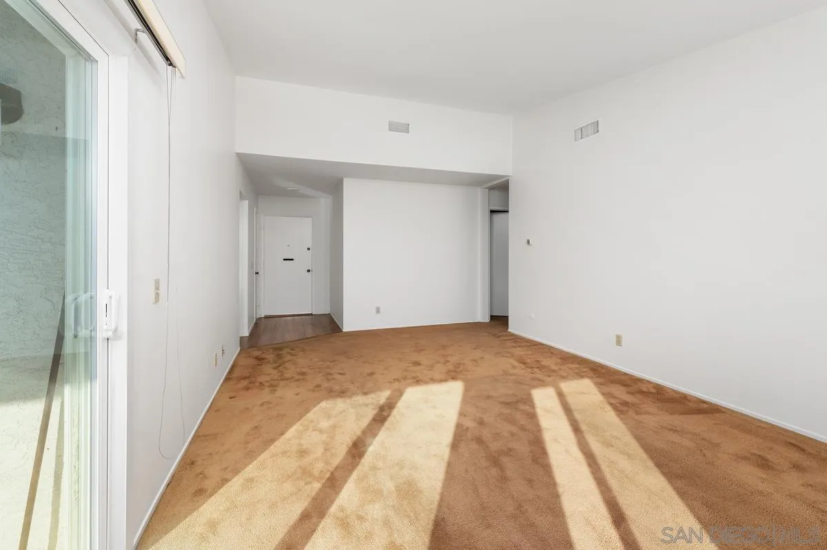 Empty carpeted living room with a sliding glass door and bright sunlight across the floor.