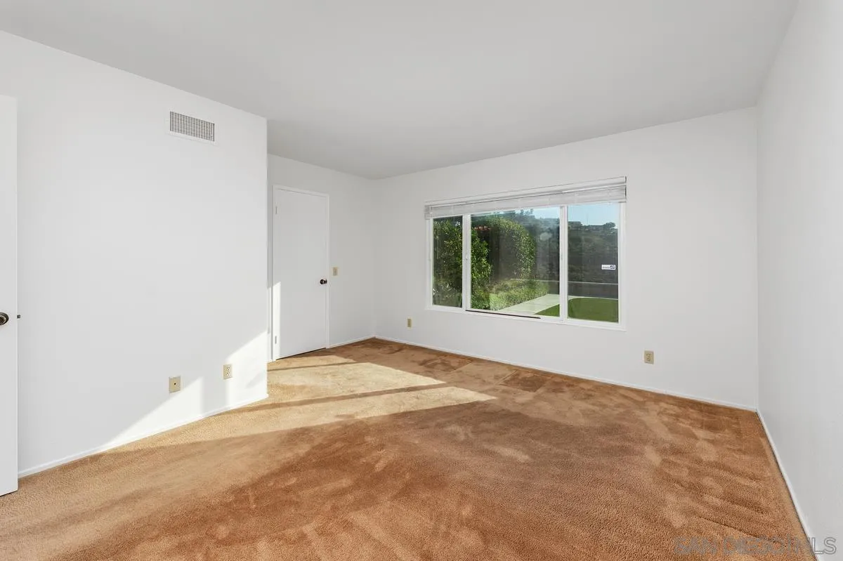 Empty carpeted room with white walls and a large window overlooking a green yard.