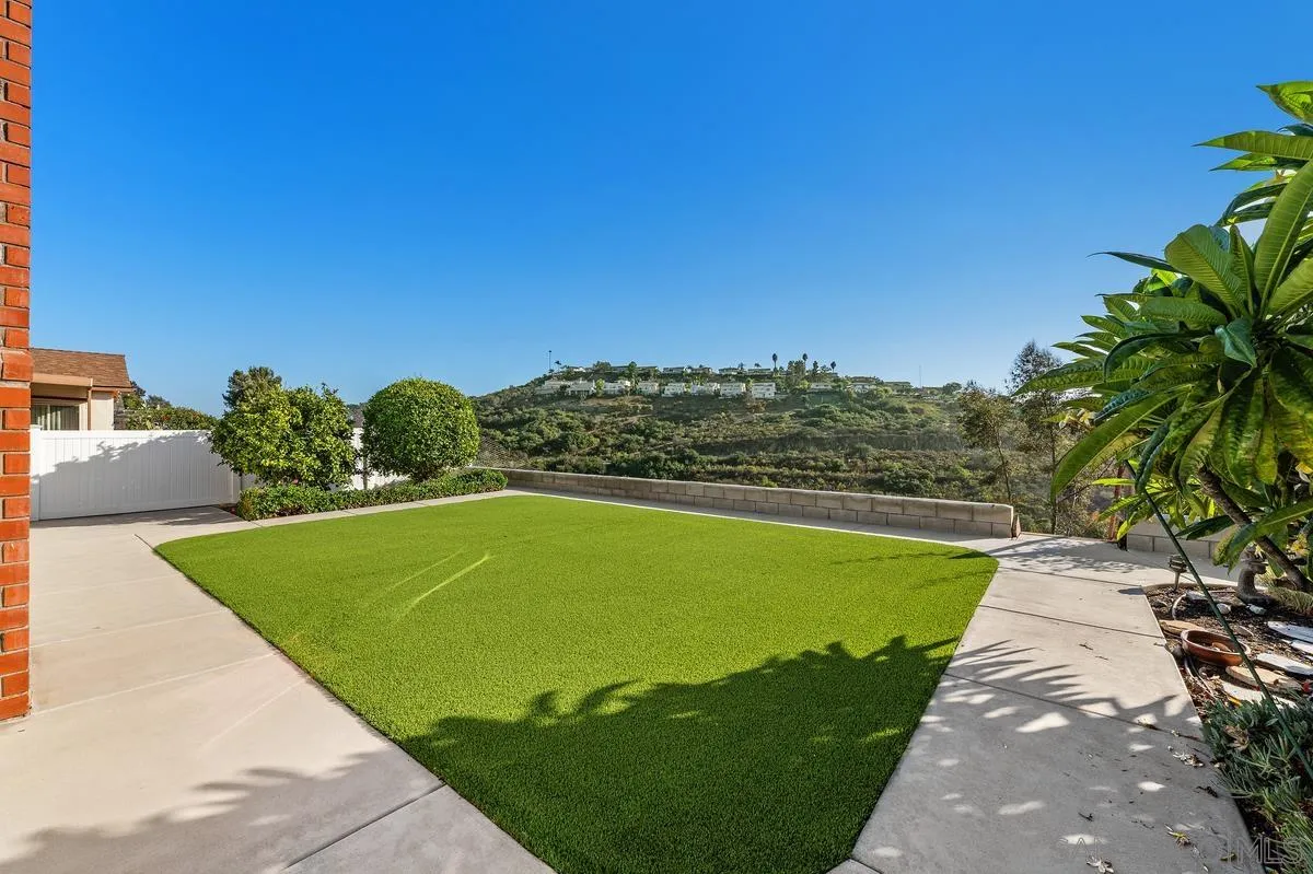 Backyard artificial-turf lawn with concrete walkways and a view of green hills.